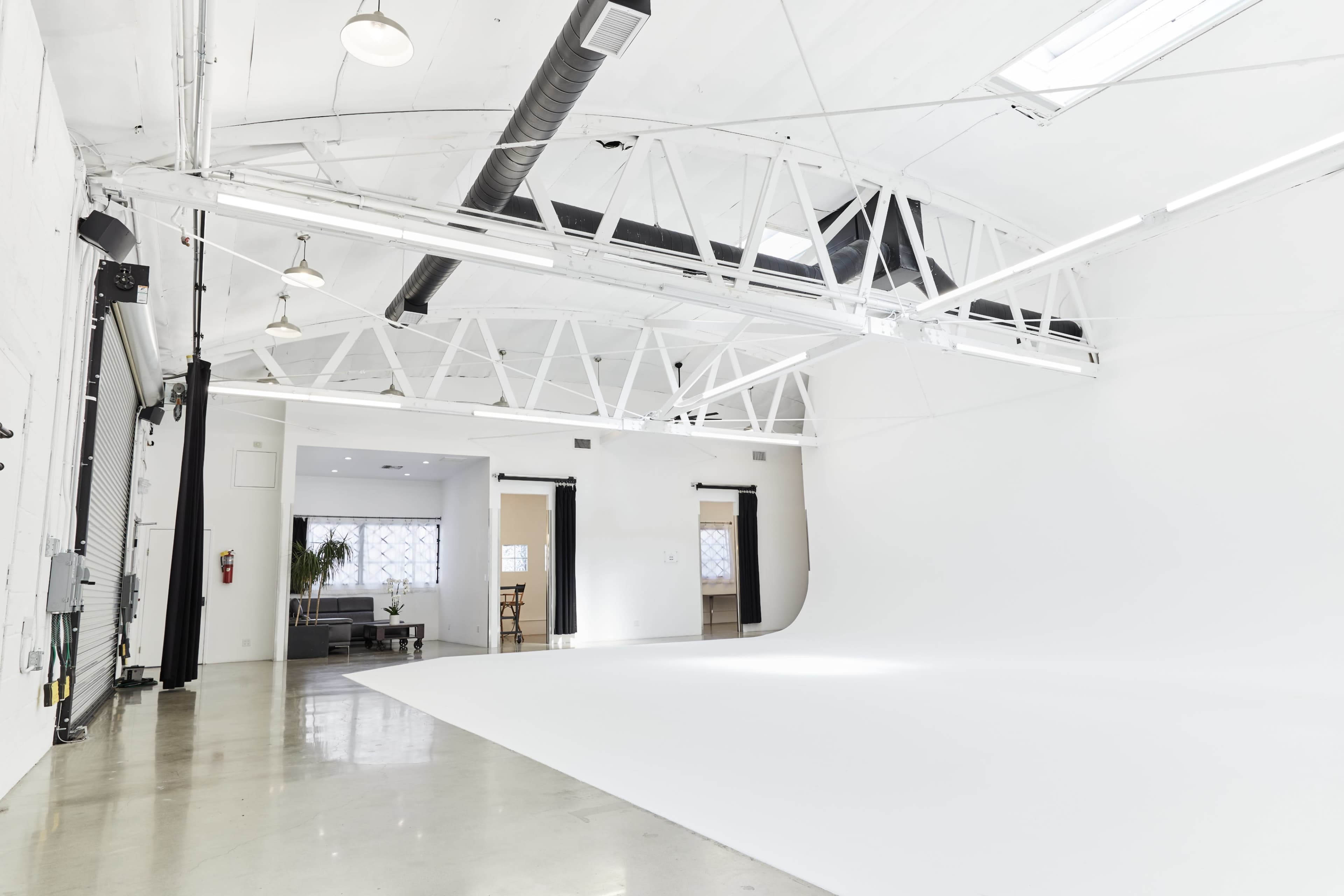 The image shows a spacious, bright studio with white walls, a seamless backdrop, and large windows allowing natural light.