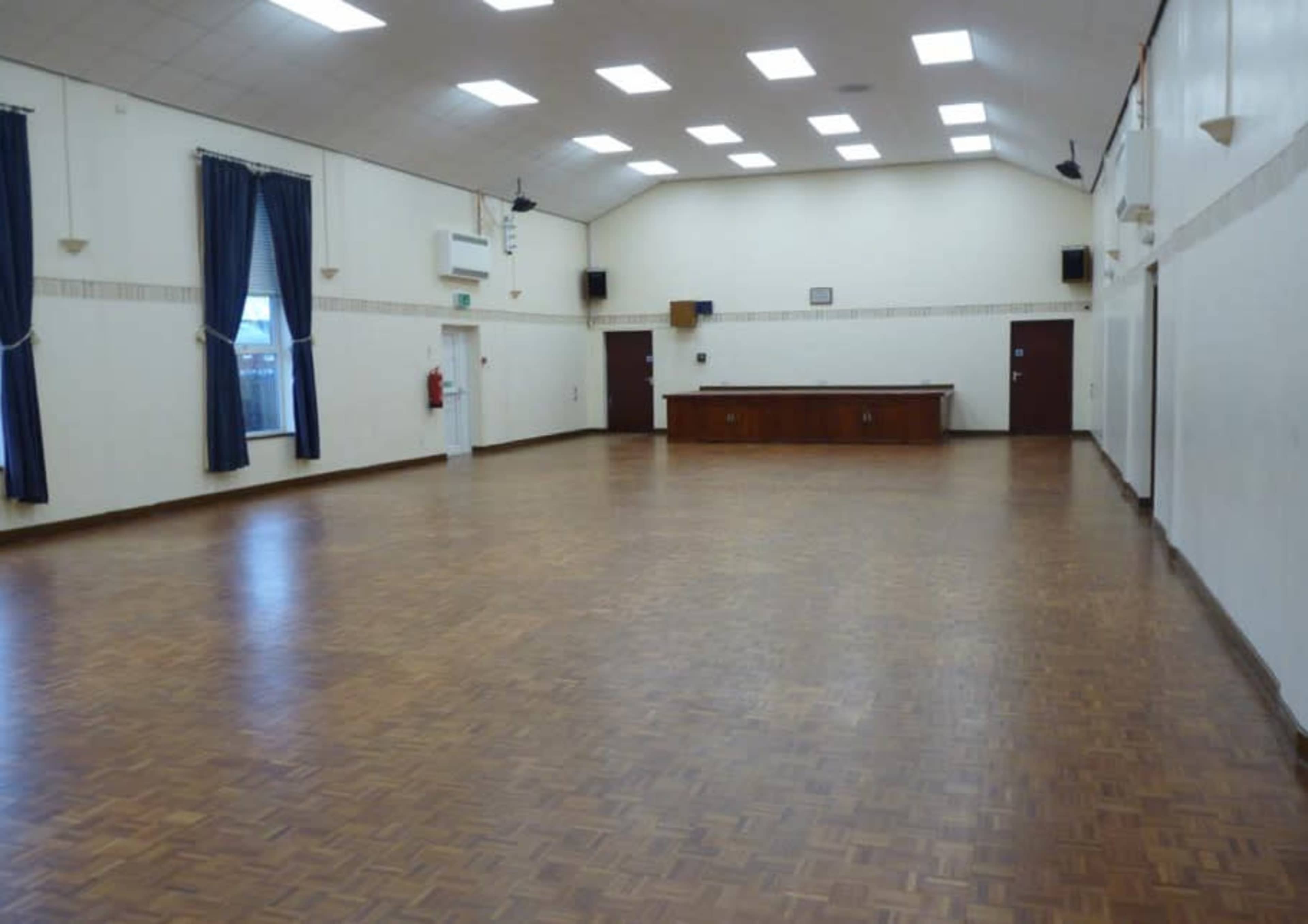 The image shows a large, empty hall with wooden flooring, ceiling lights, and a service counter at the far end.