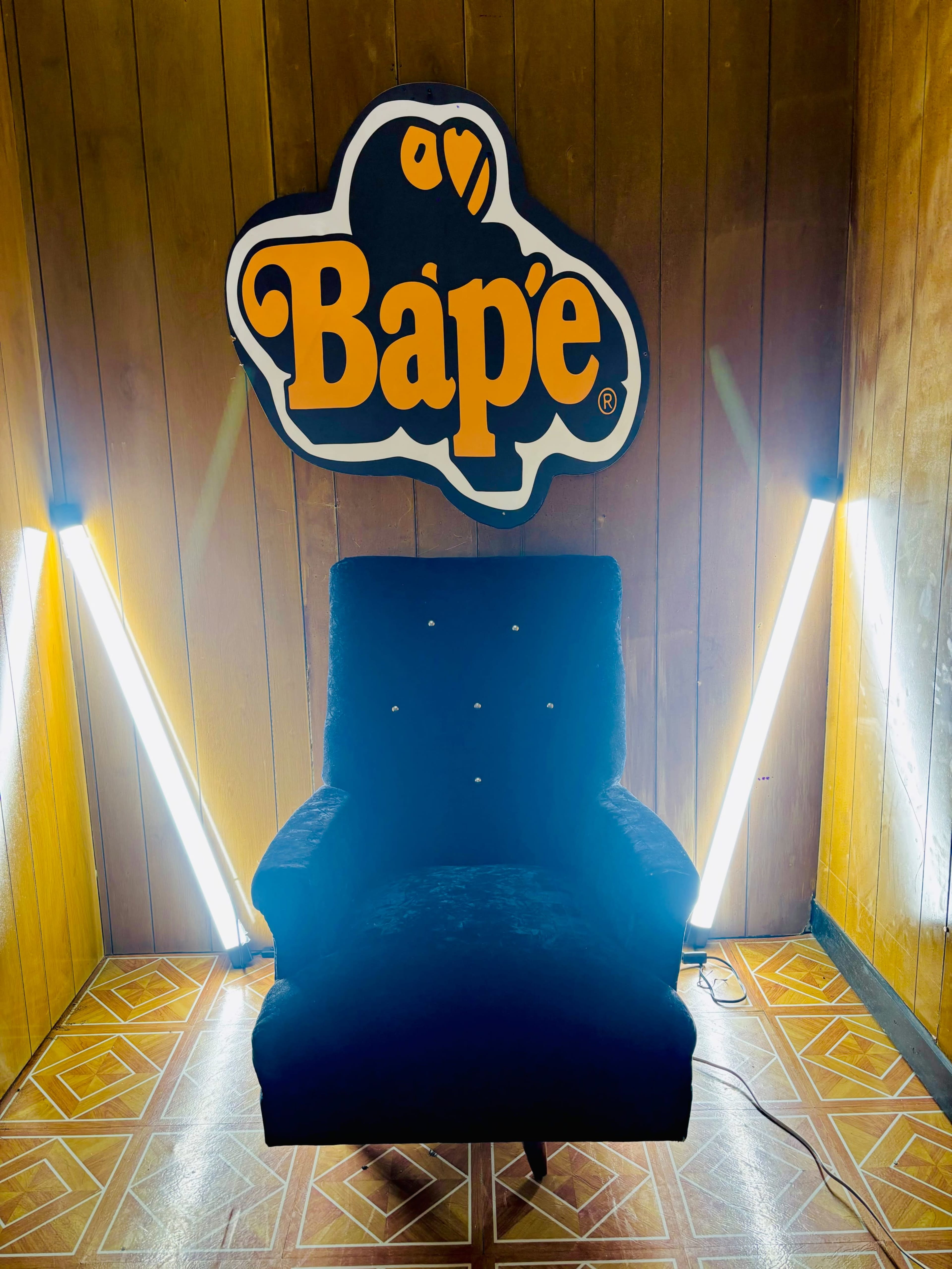 A black velvet chair is positioned in front of a large Bape logo on a wooden wall, illuminated by two bright light fixtures.