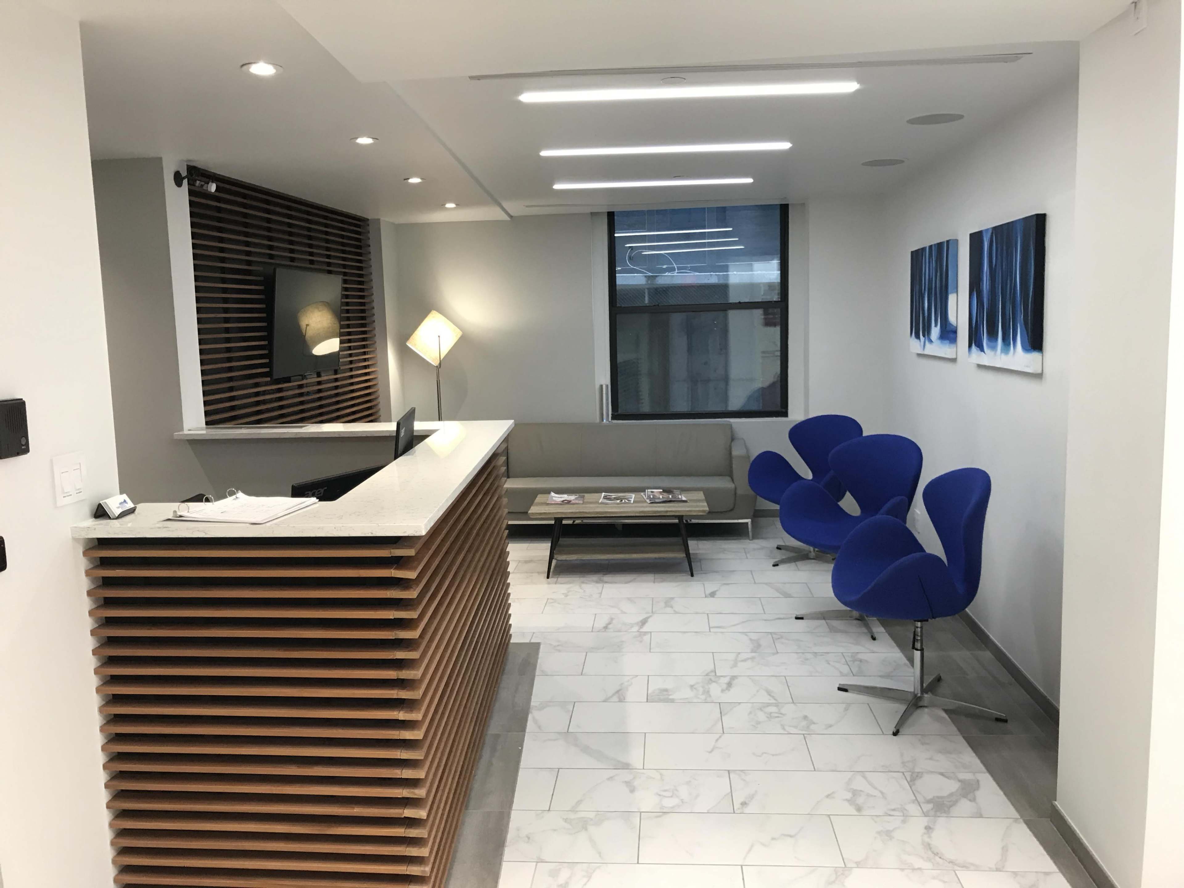 The image shows a modern office reception area with a wooden front desk, seating area featuring blue chairs, and a coffee table on a marble floor.