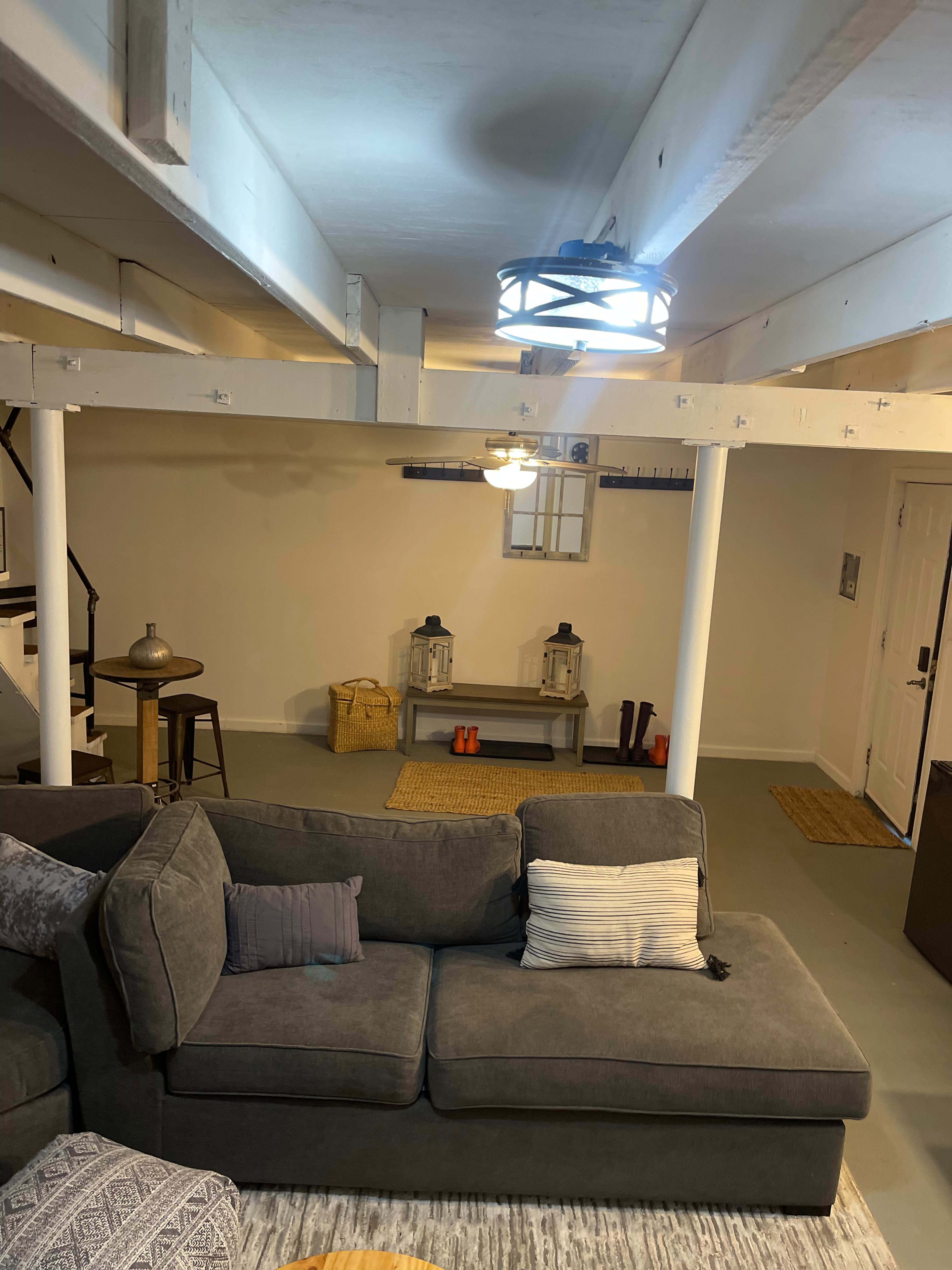 A well-lit basement with a gray sectional sofa, a small dining table, and decorative lanterns on a console table against a white wall.