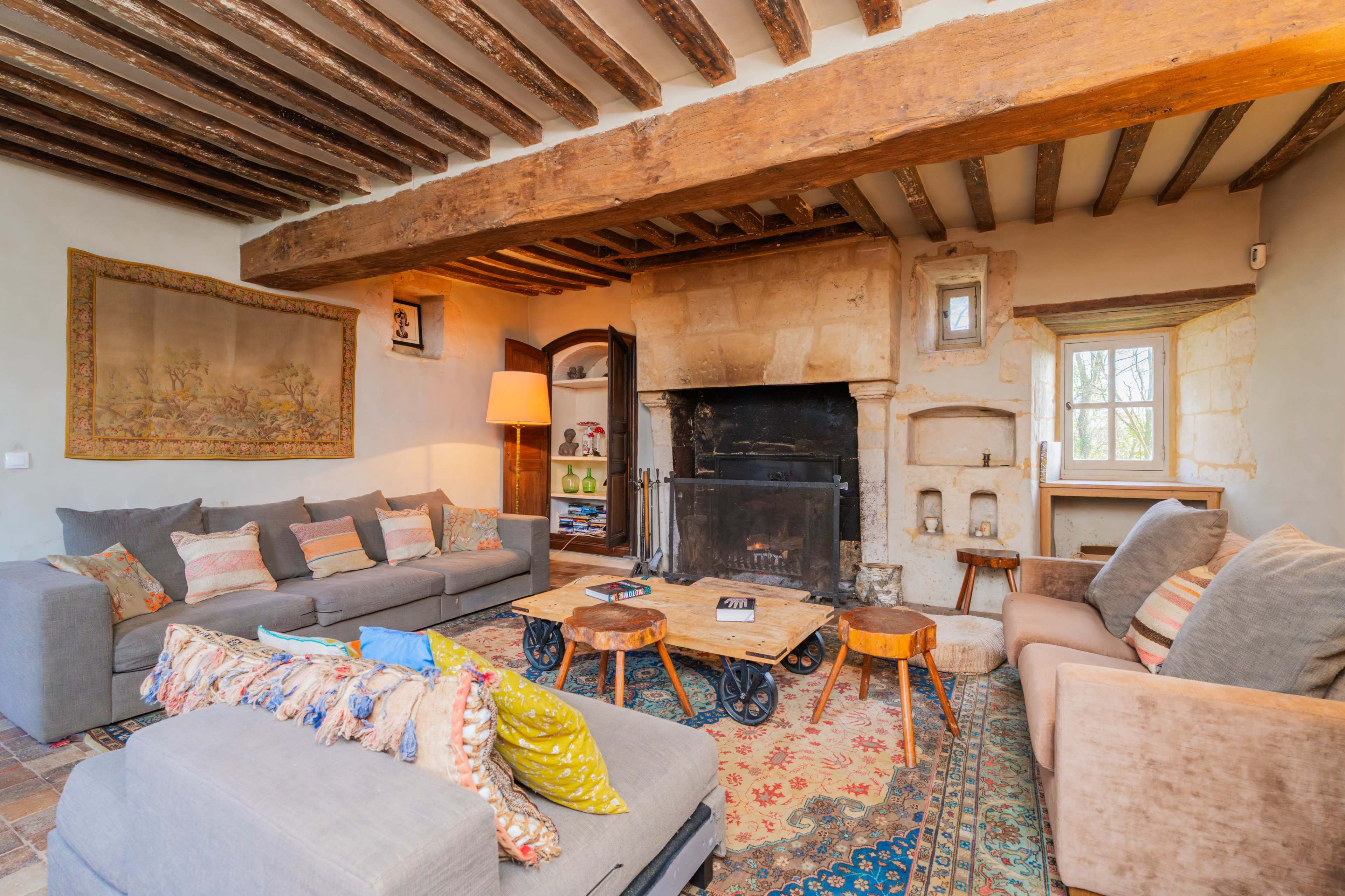 The image shows a cozy living room with exposed wooden beams, a stone fireplace, and various seating arrangements including gray sofas and wooden coffee tables.