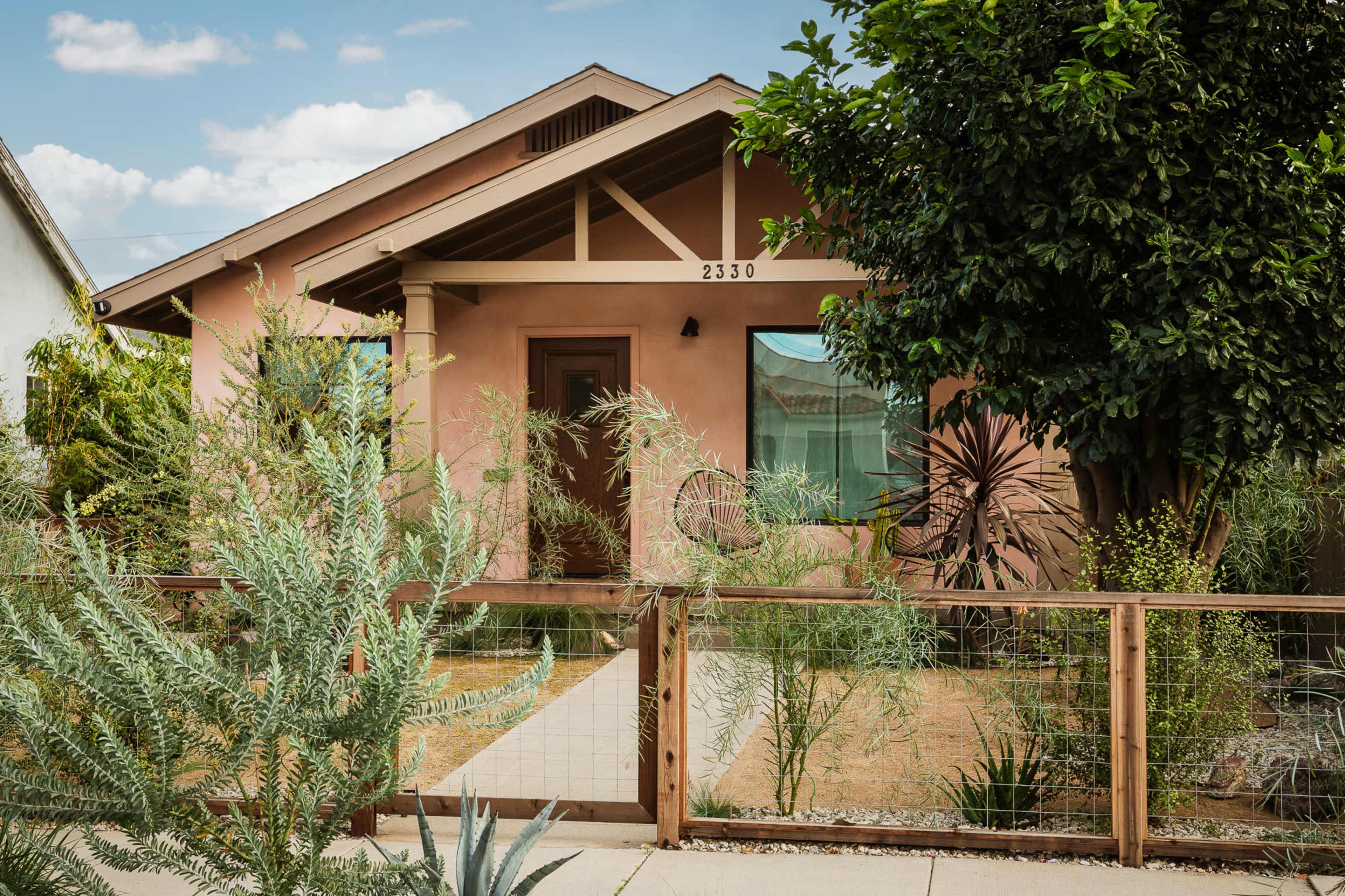 The image shows a single-story house with a light brown exterior and a wooden fence surrounding a garden with various plants.
