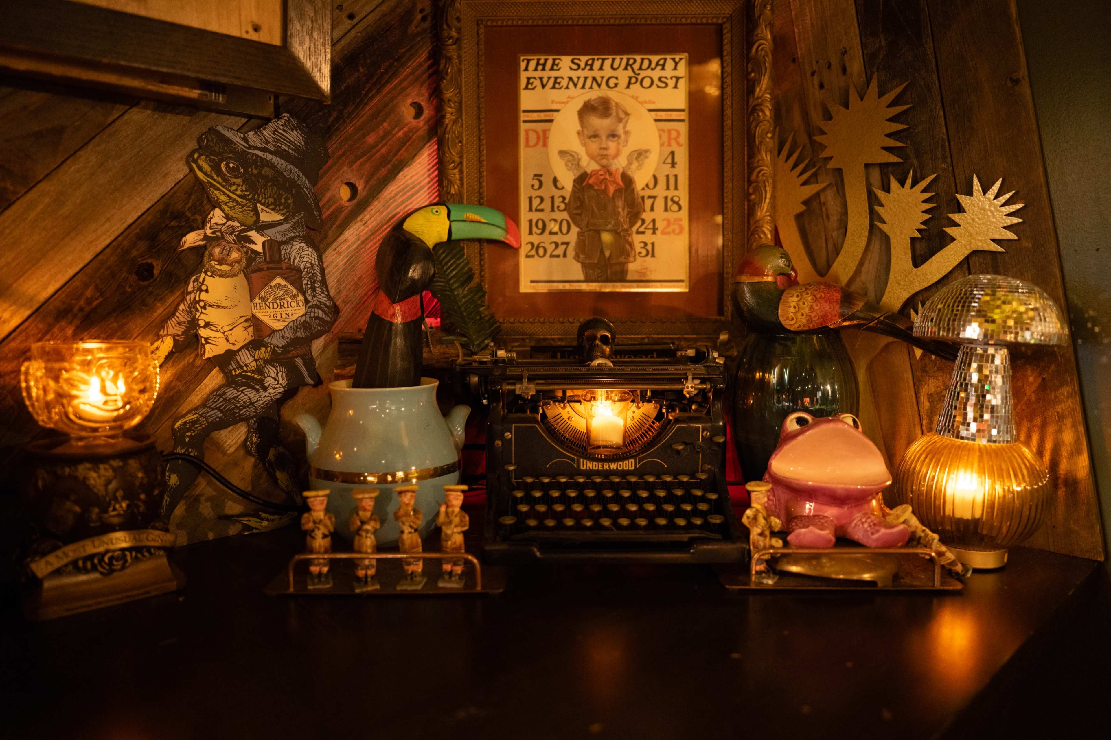 The image features a vintage decor setup with a typewriter, a framed "Saturday Evening Post" cover, and various eclectic figurines and lights arranged on a dark wooden surface.