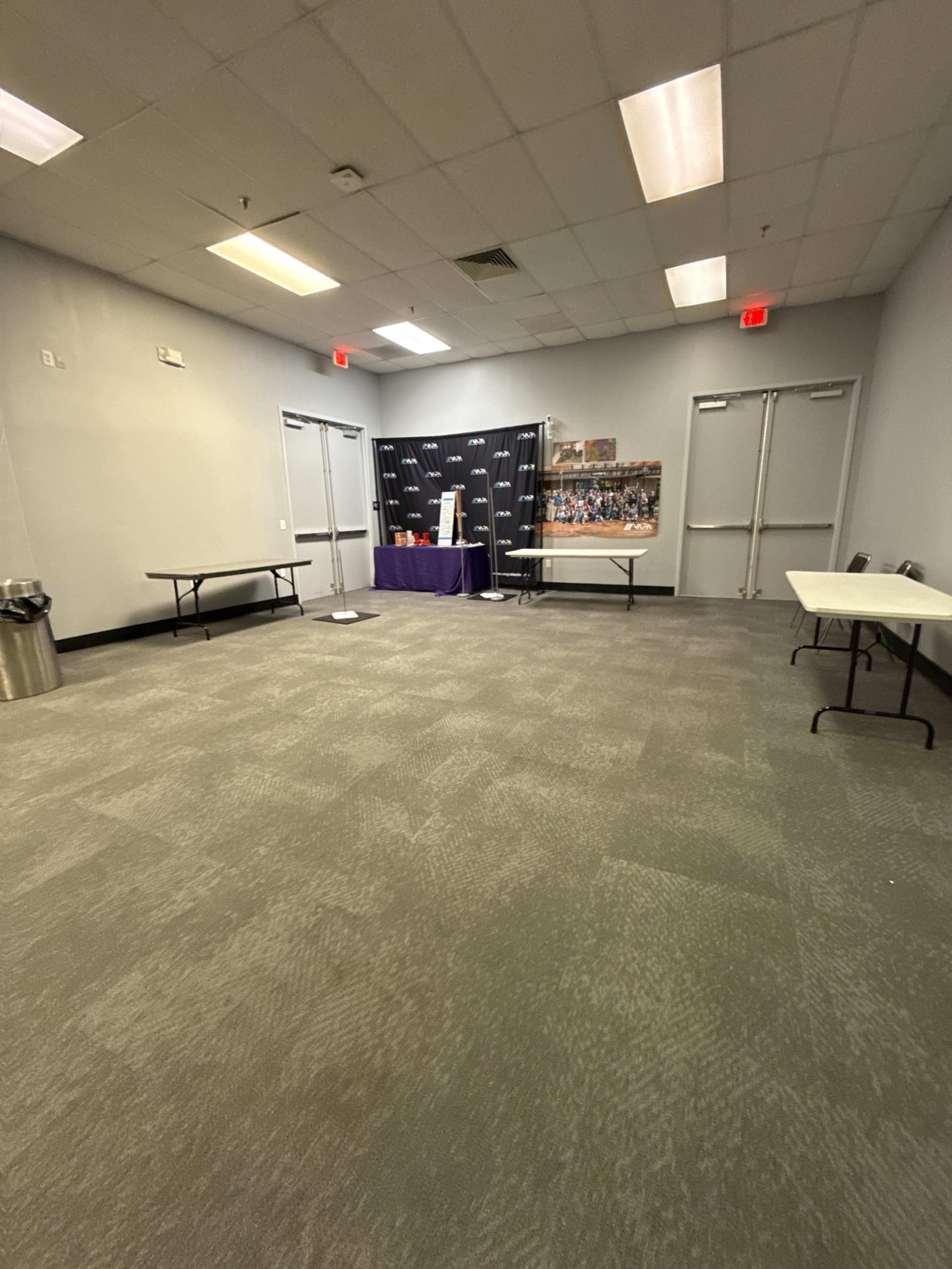 The image shows a sparsely furnished room with two tables and a backdrop featuring a black cloth, along with double doors and a trash can in the corner.