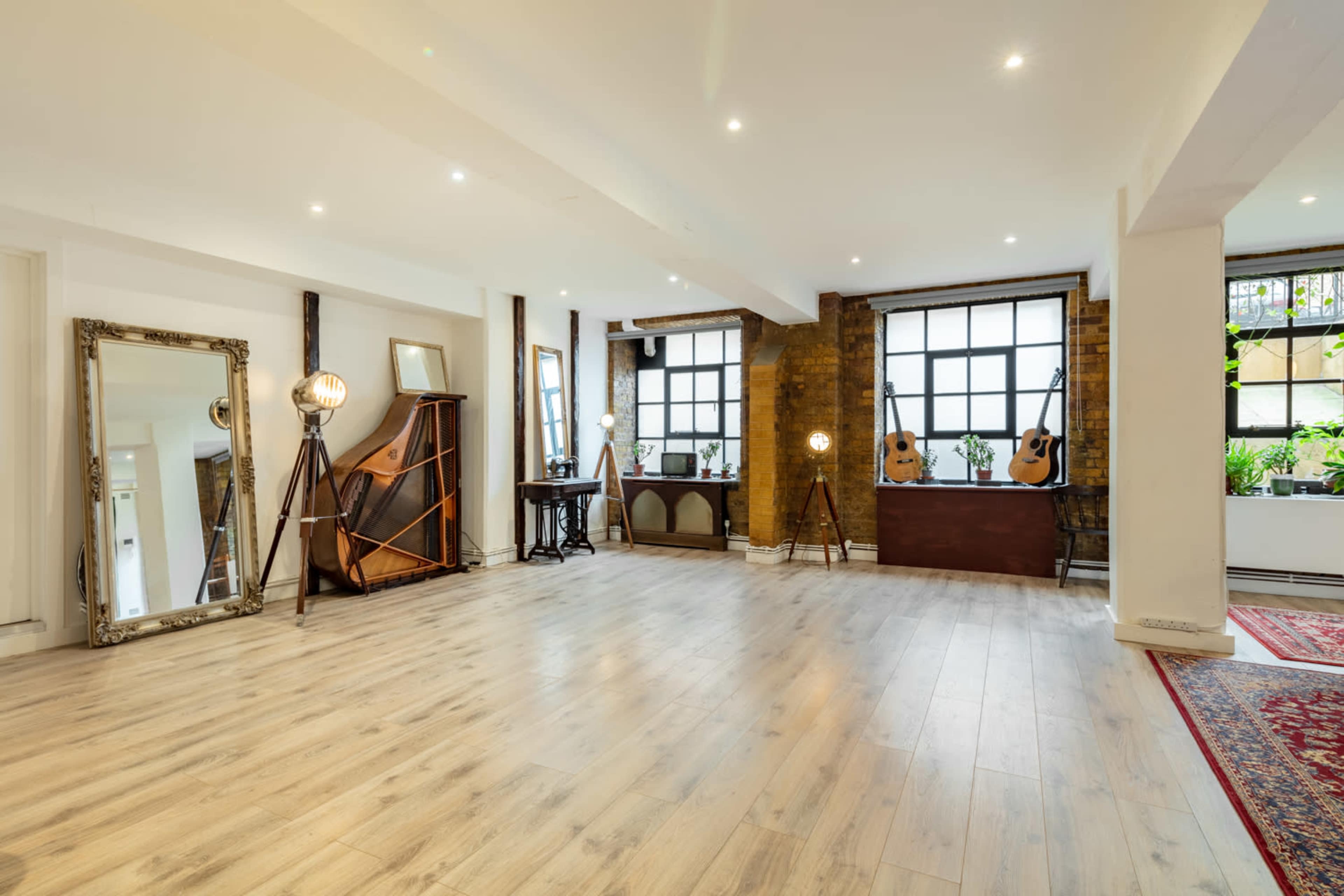 A spacious room features wooden flooring, large windows, and a mix of decorative furniture, including mirrors and guitars.