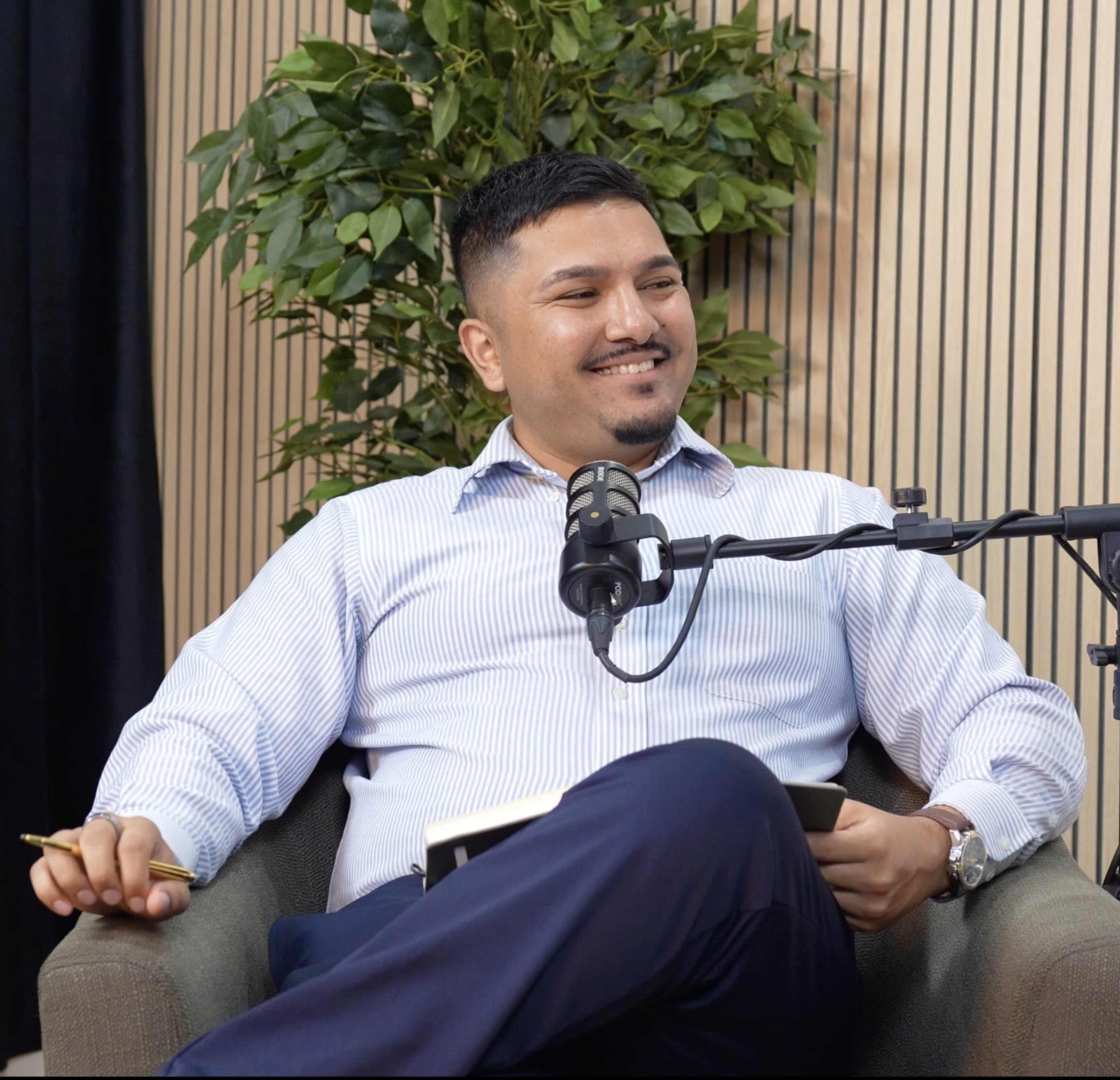 A man in a striped shirt sits comfortably in a chair, holding a pen, while engaging in a conversation near a microphone and a potted plant.