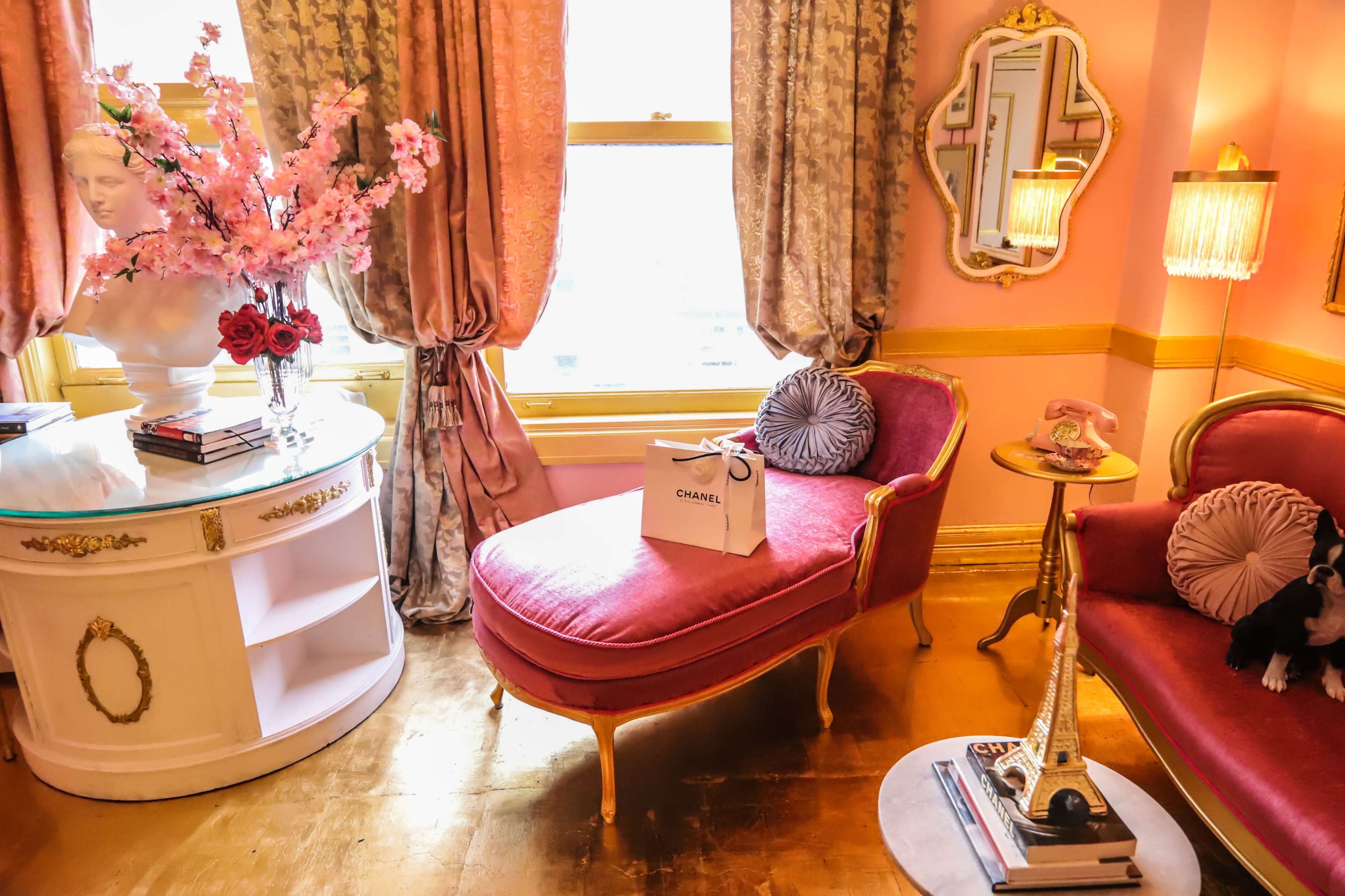 A pink-themed sitting room features a chaise lounge, a small table with an Eiffel Tower figurine, and floral decorations.