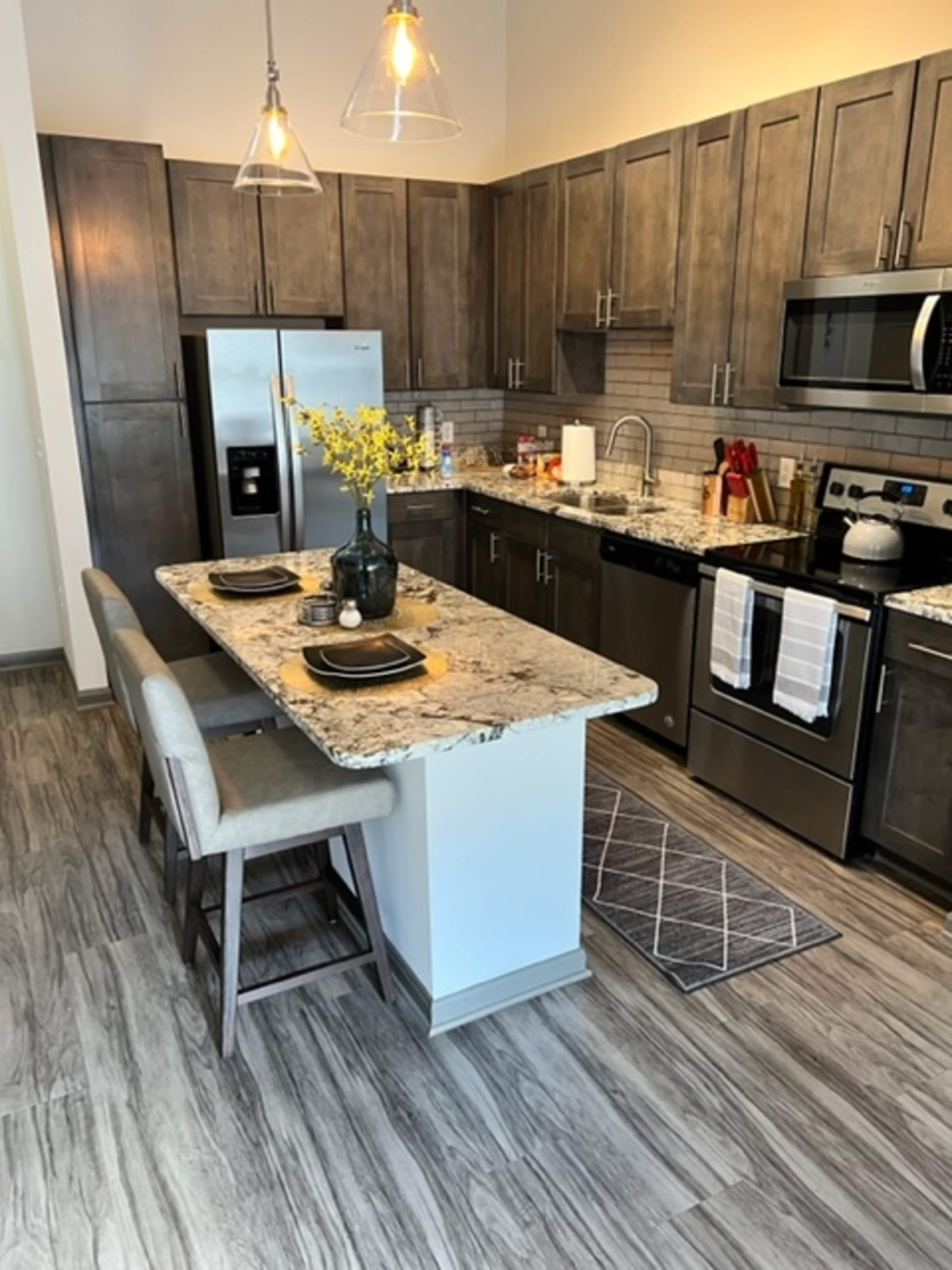 The image shows a modern kitchen with wooden cabinets, stainless steel appliances, a granite countertop, and a small island with seating.
