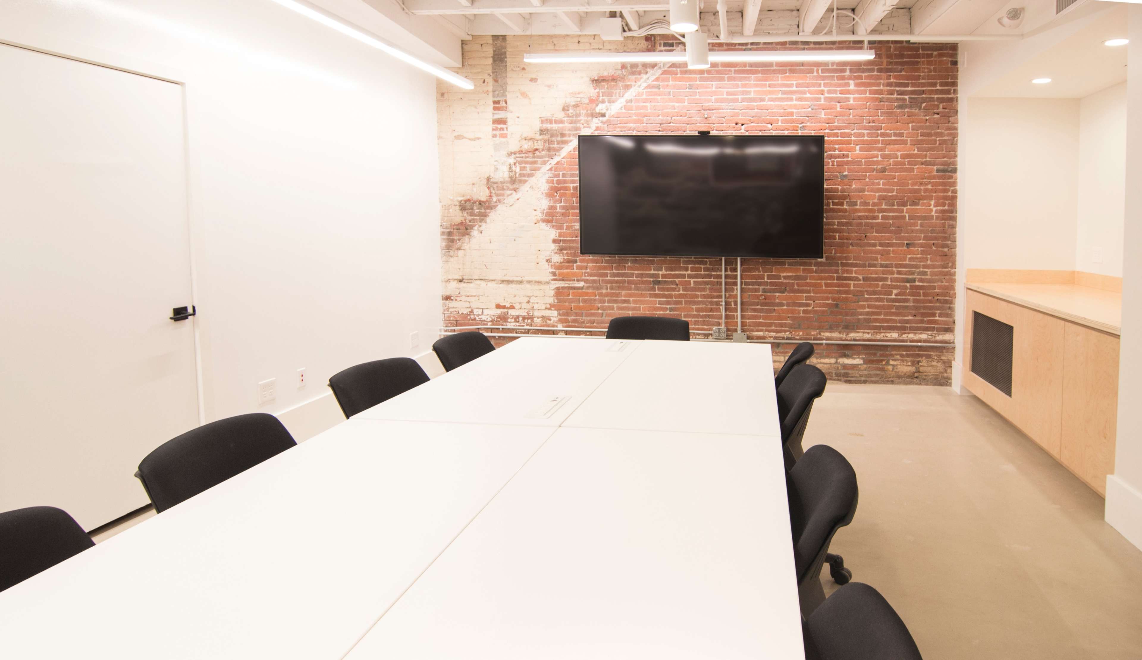 A modern conference room features a long white table surrounded by black chairs, with a large television mounted on a brick wall.