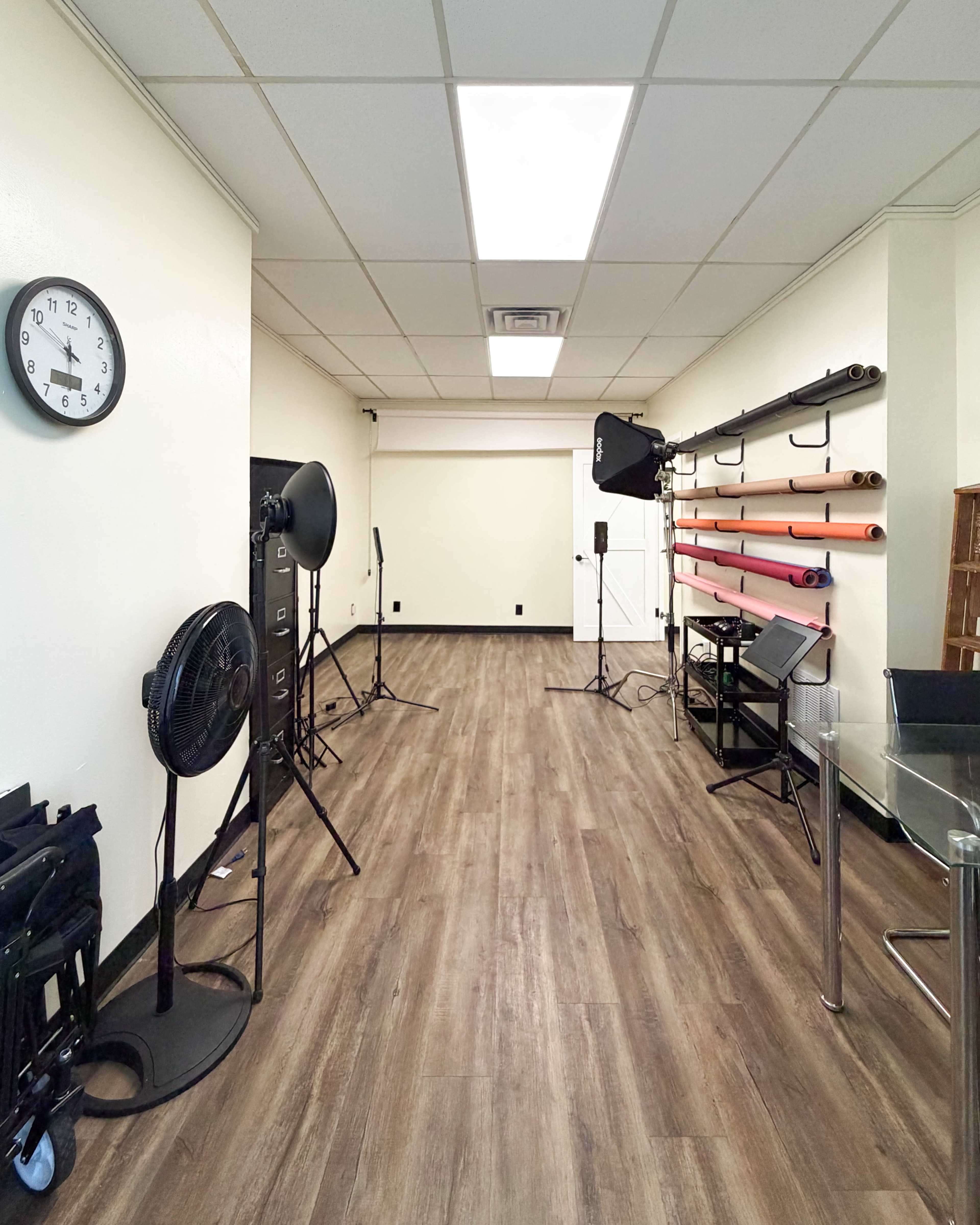 The image shows a spacious photography studio with wooden flooring, a clock on the wall, lighting equipment, and colorful backdrops neatly arranged along one side.