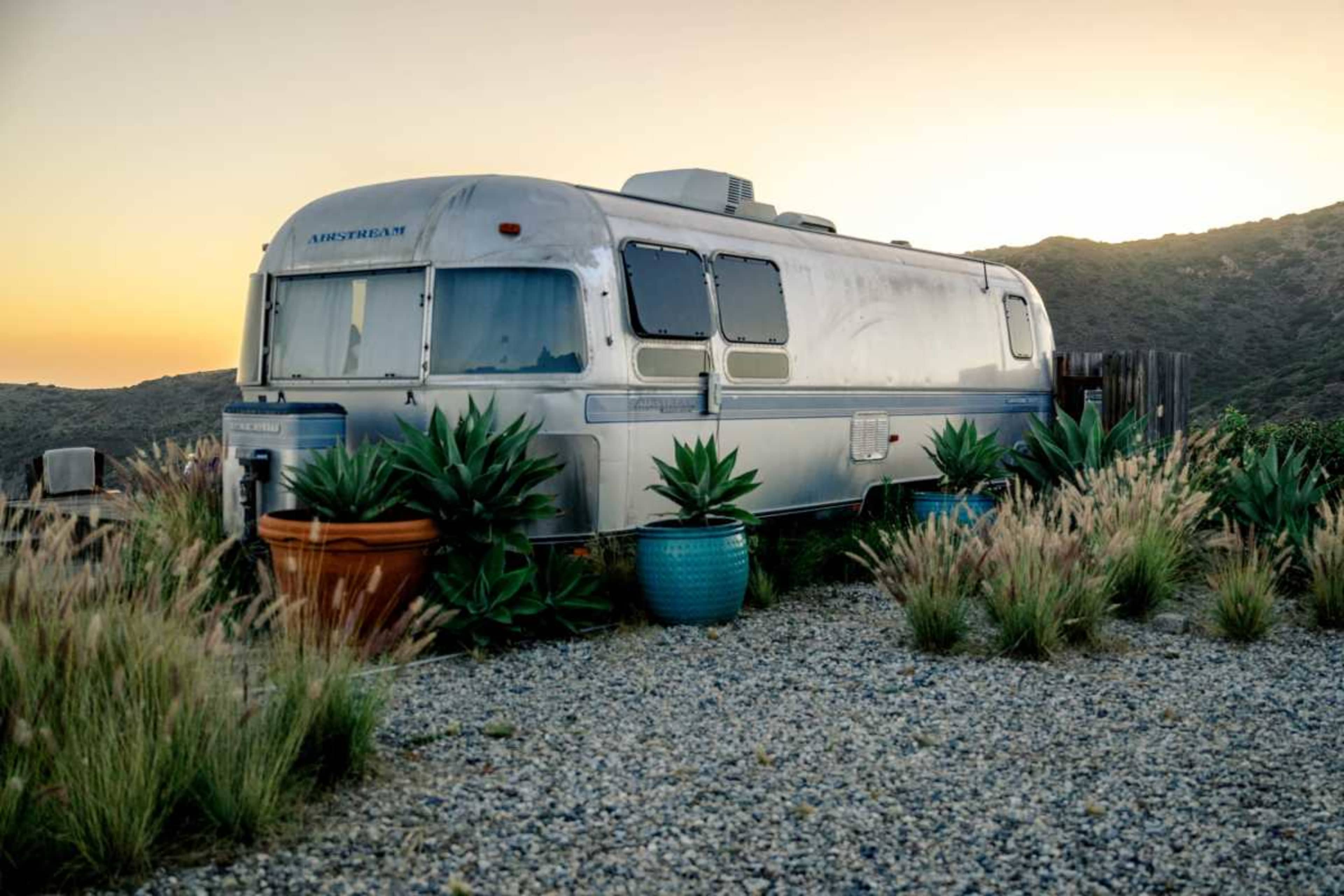 An Airstream trailer is surrounded by potted plants and gravel with mountains in the background at sunset.