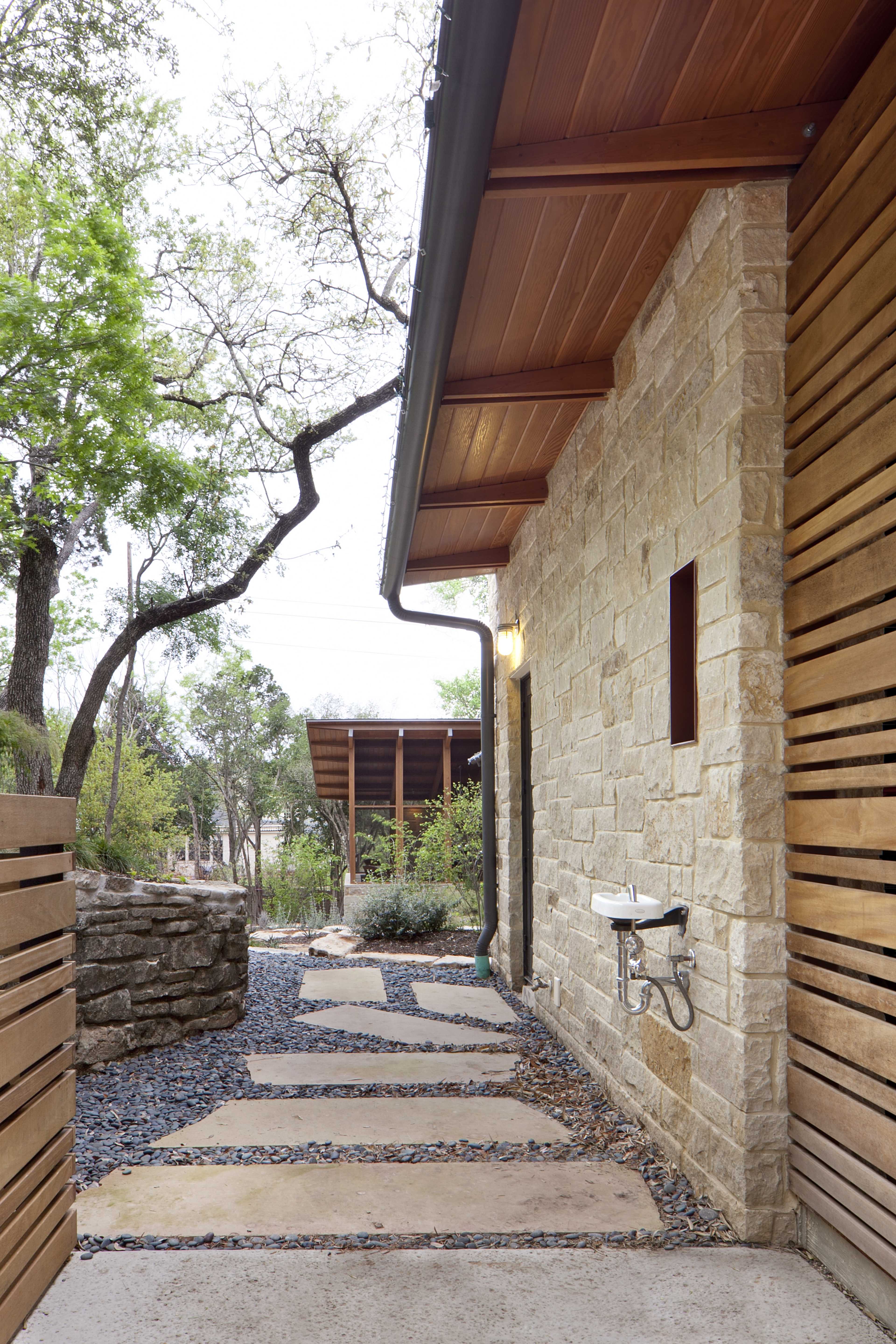The image shows a stone pathway bordered by landscaped greenery, leading toward a building with wooden siding and a covered area in the background.
