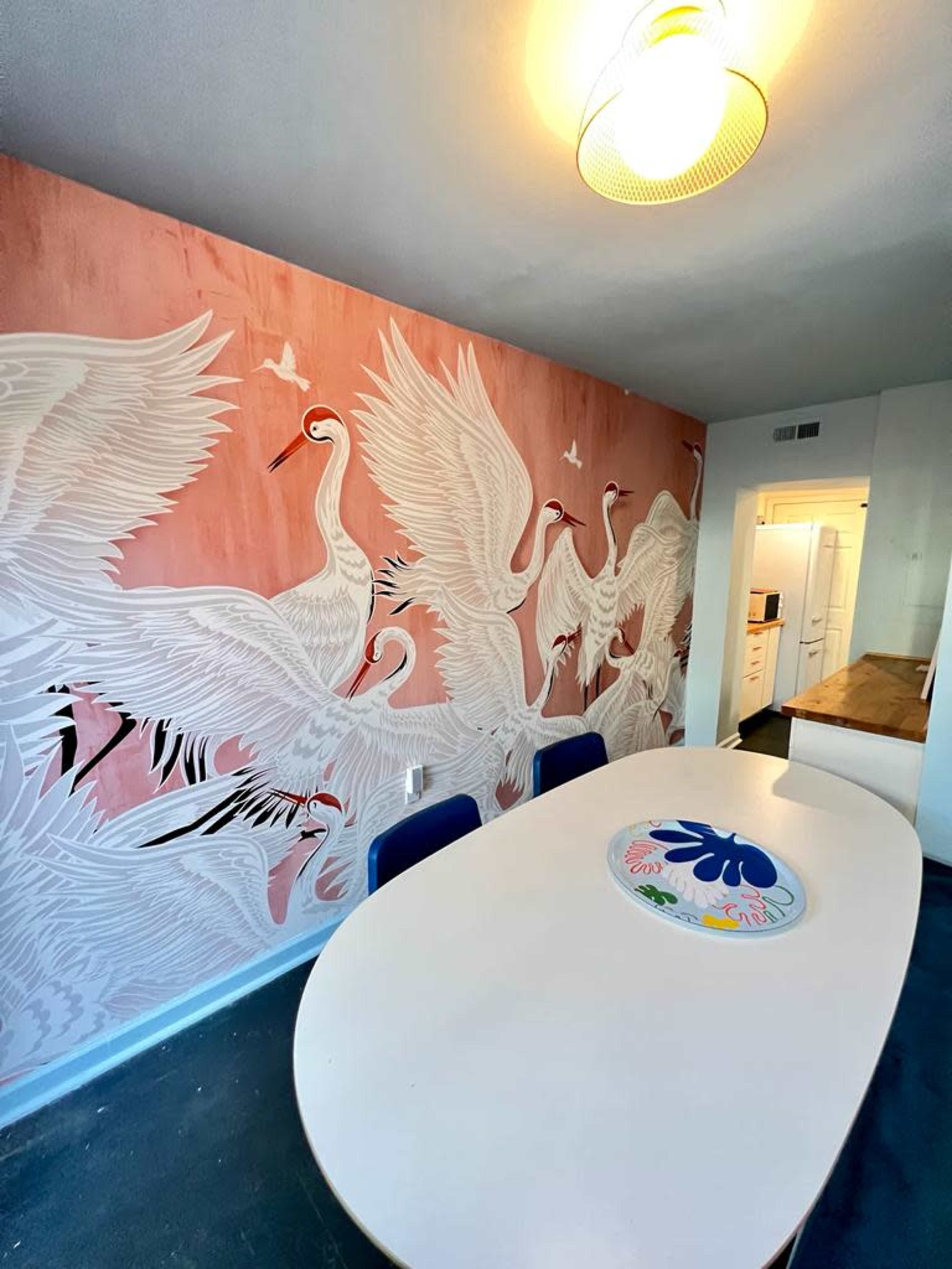 A dining area with a large oval table surrounded by chairs, set against a pink wall adorned with a mural of white cranes in flight.