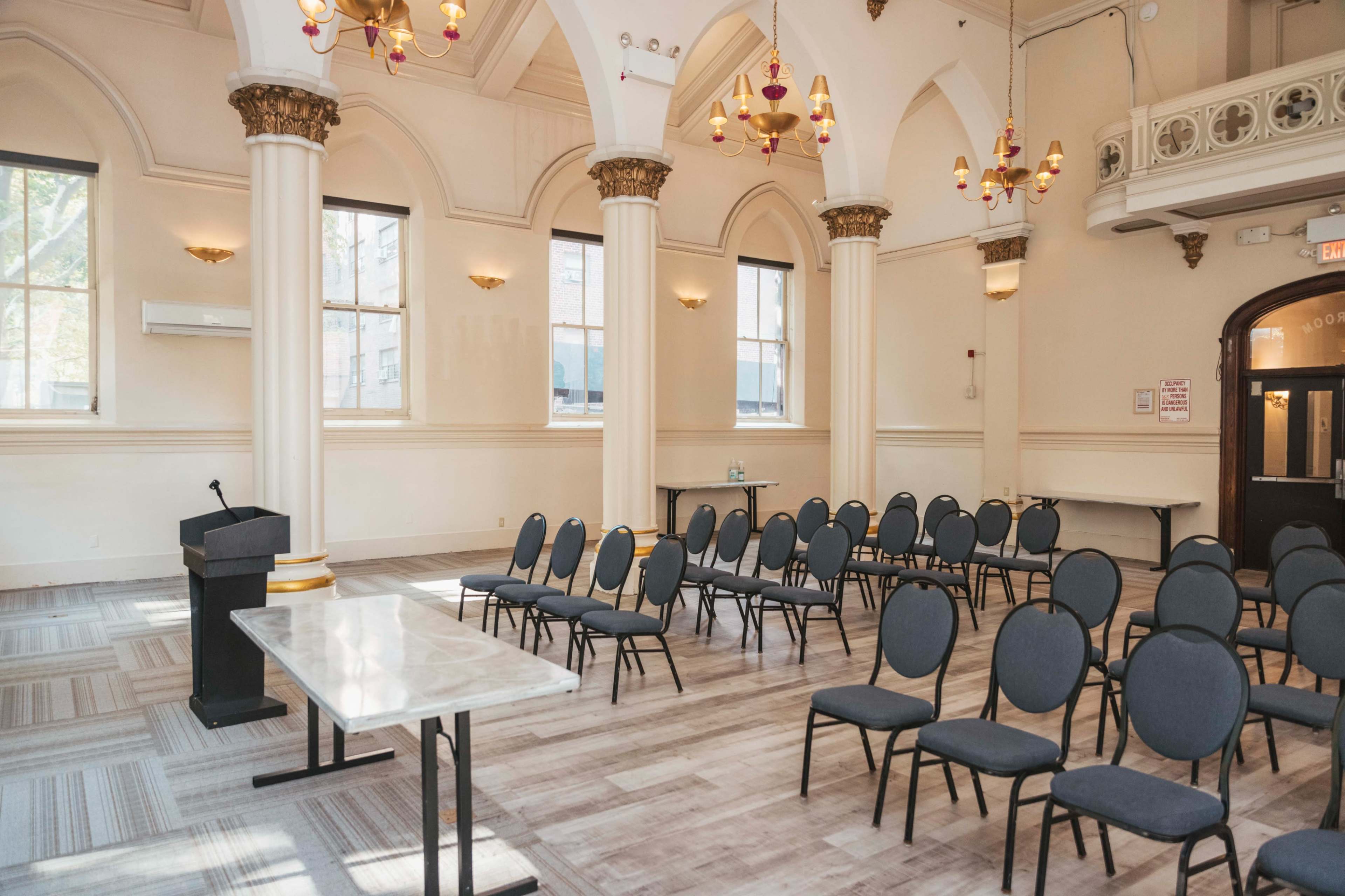 The image shows a spacious event room with arranged chairs facing a podium and a table, featuring high ceilings adorned with decorative lighting and architectural details.