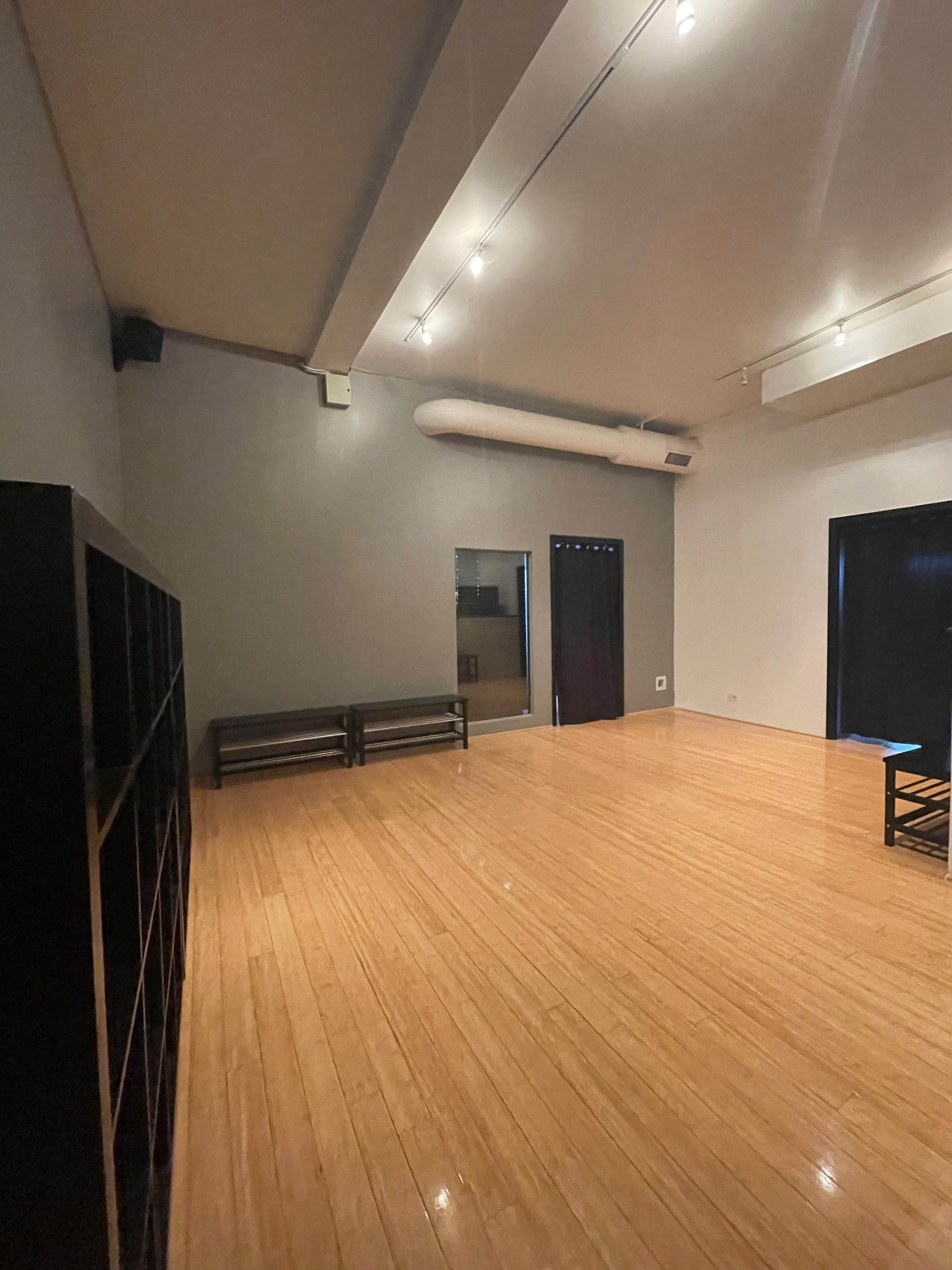 A minimalistic interior room with wooden flooring and sparse furniture, featuring a bench against one wall and a large window with dark curtains.