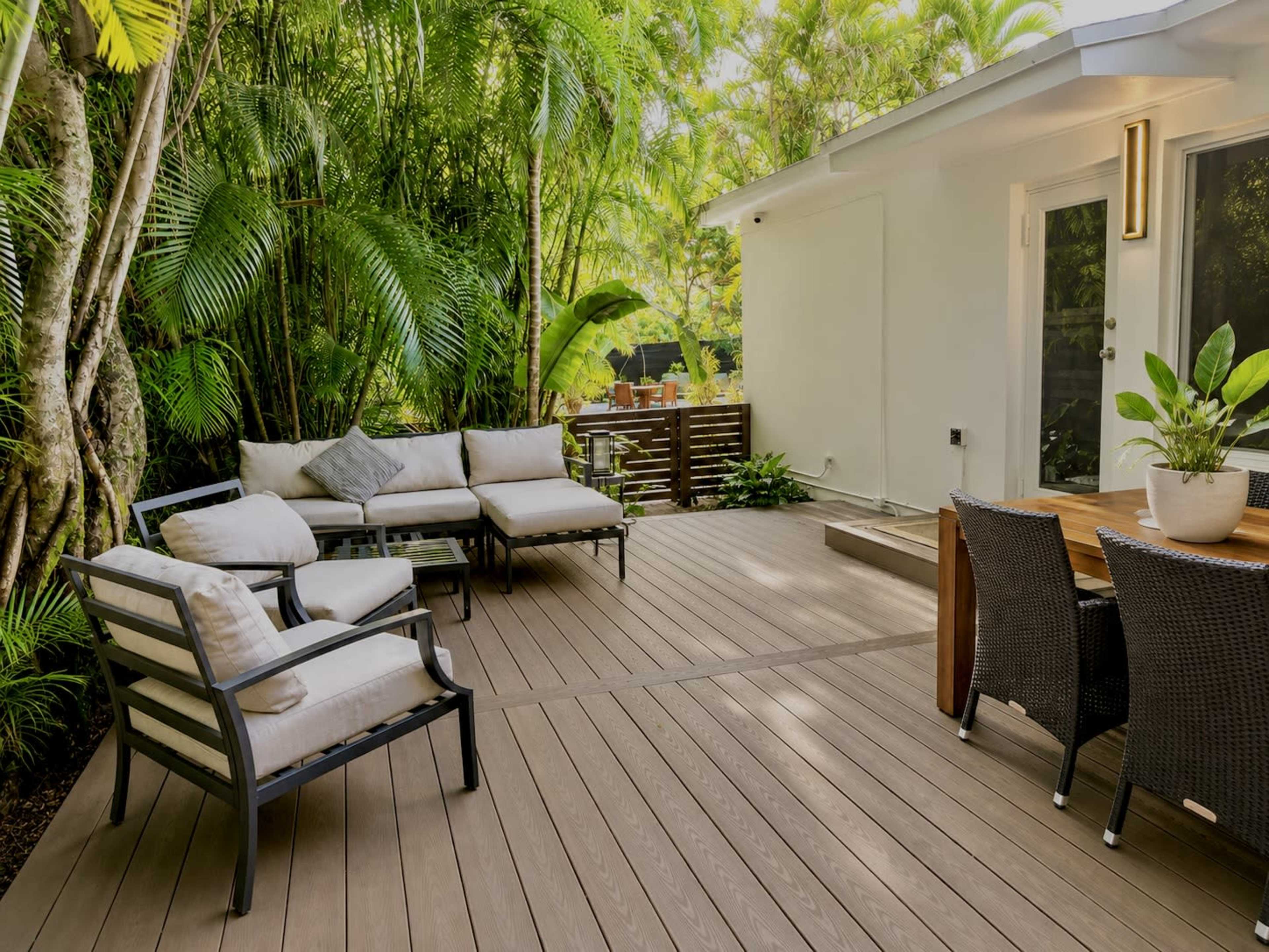 The image shows a patio area with seating on a wooden deck surrounded by lush green tropical plants.