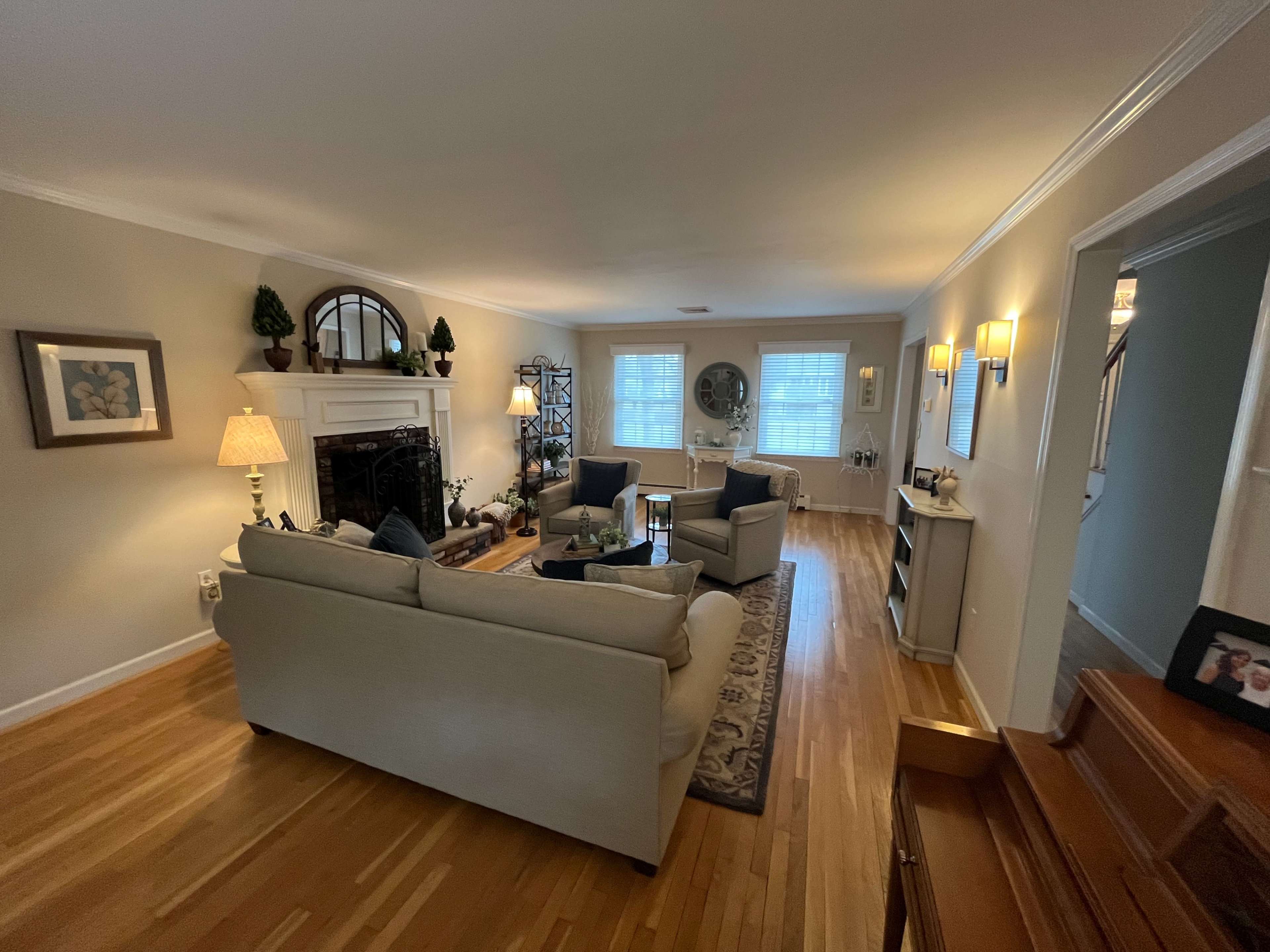 The living room features a light-colored couch, two armchairs, a fireplace, and windows allowing natural light to enhance the wood flooring.