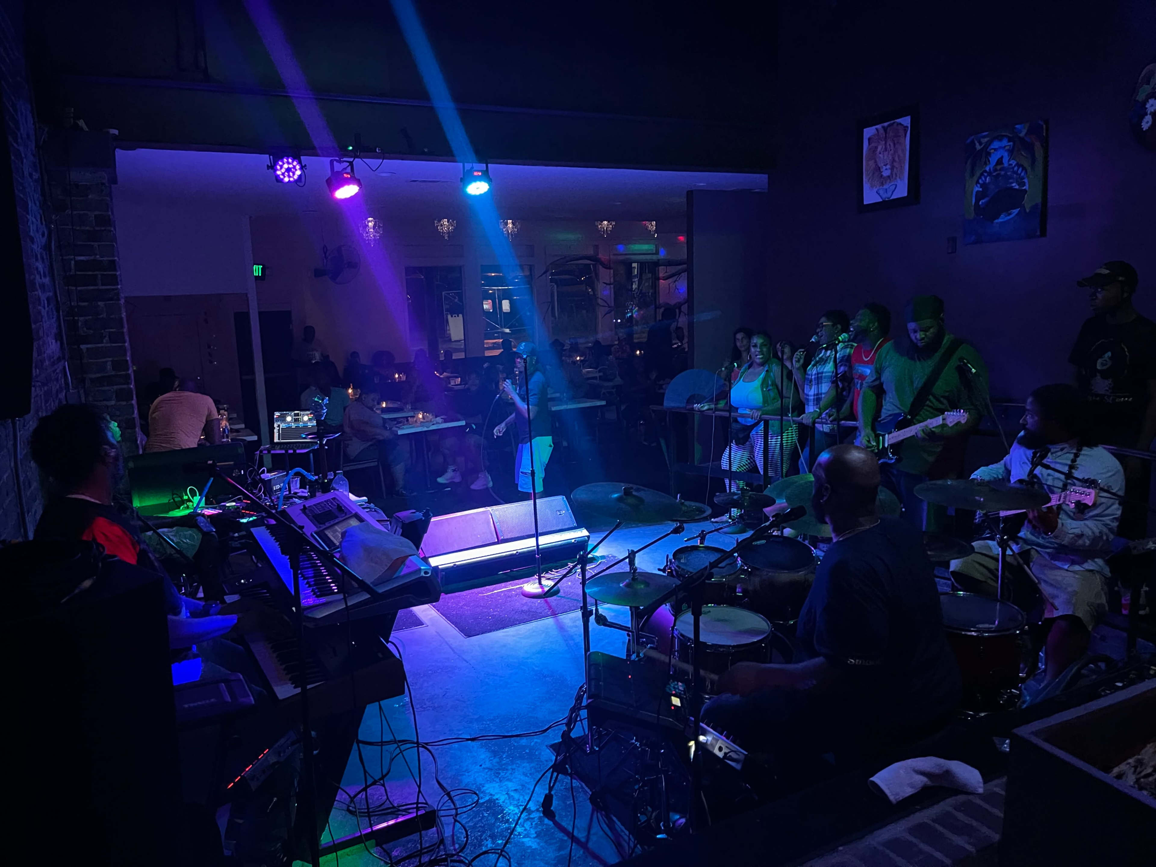 A band performs on stage in a dimly lit venue while a small audience watches from the side.