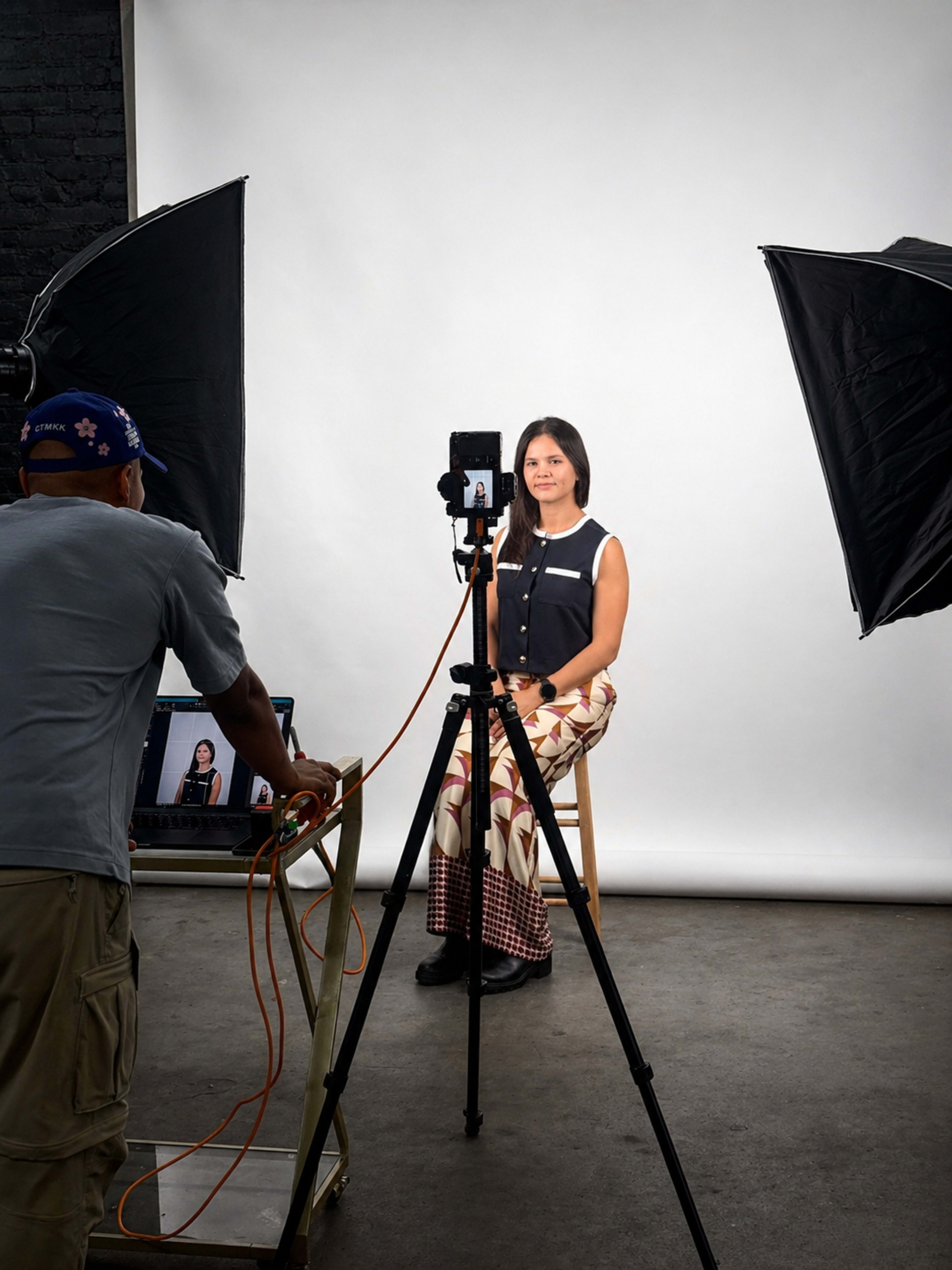 A person is sitting on a stool in a studio setting, while a photographer adjusts the camera and lighting equipment.