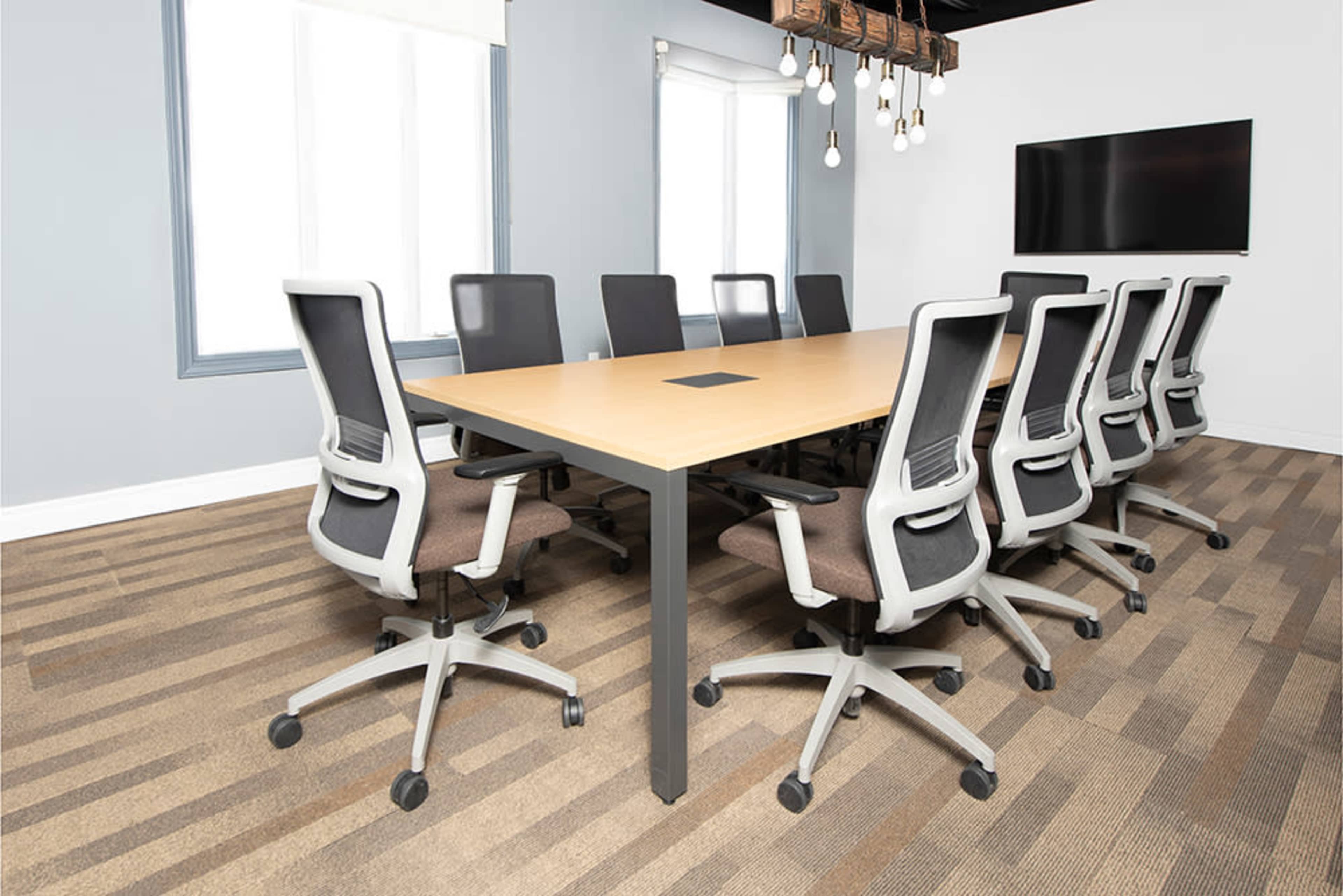 A long conference table with gray office chairs is arranged in a well-lit meeting room featuring a television on the wall.