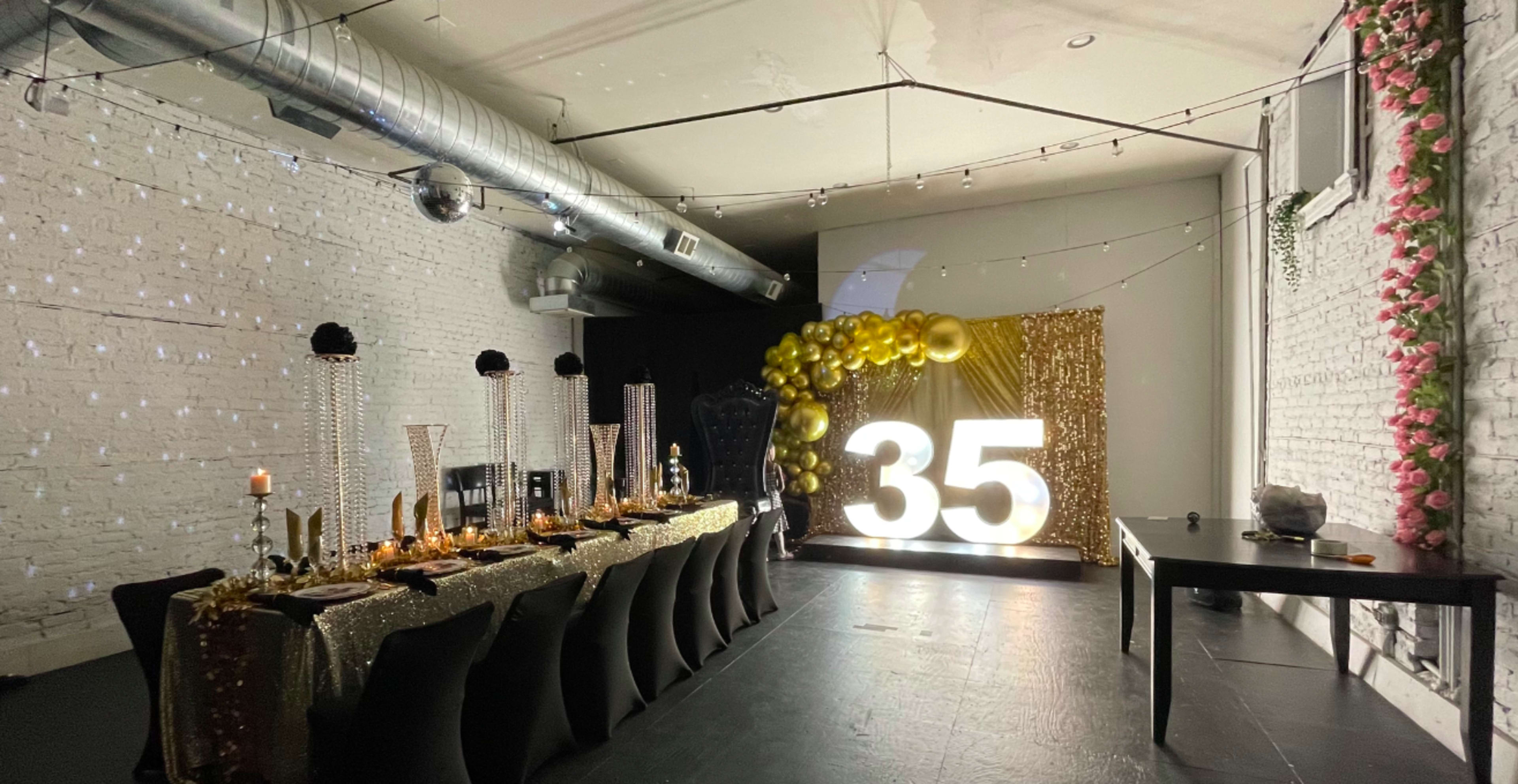 A decorated space features a long table set for a celebration, with a large illuminated "35" and golden balloon decor in the background.