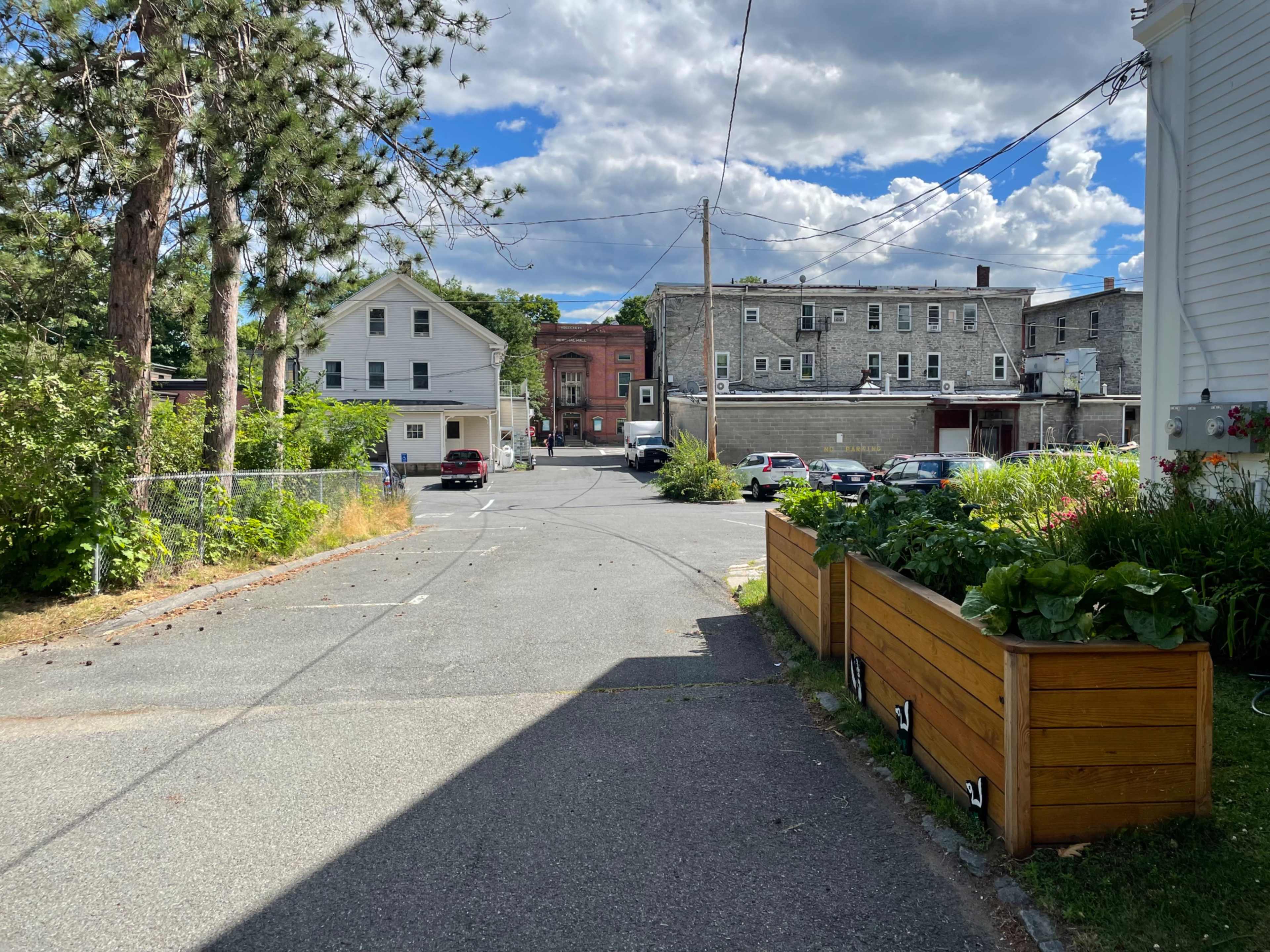 A narrow alleyway is lined with greenery and leads to a gravel road, with several parked cars and buildings in the background under a partly cloudy sky.