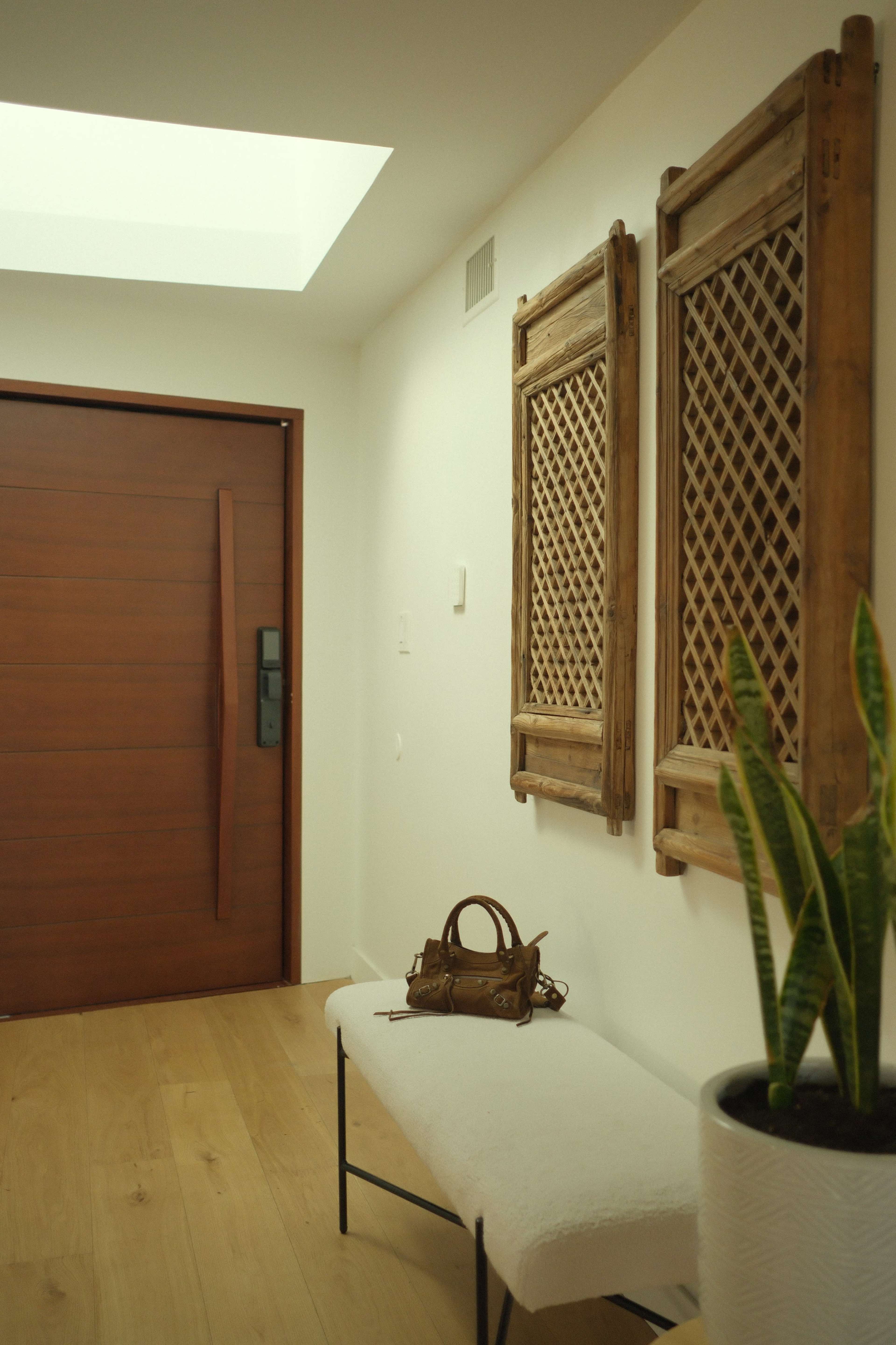 A modern entryway features a wooden door, two decorative wall panels, a bench, a purse, and a potted plant.