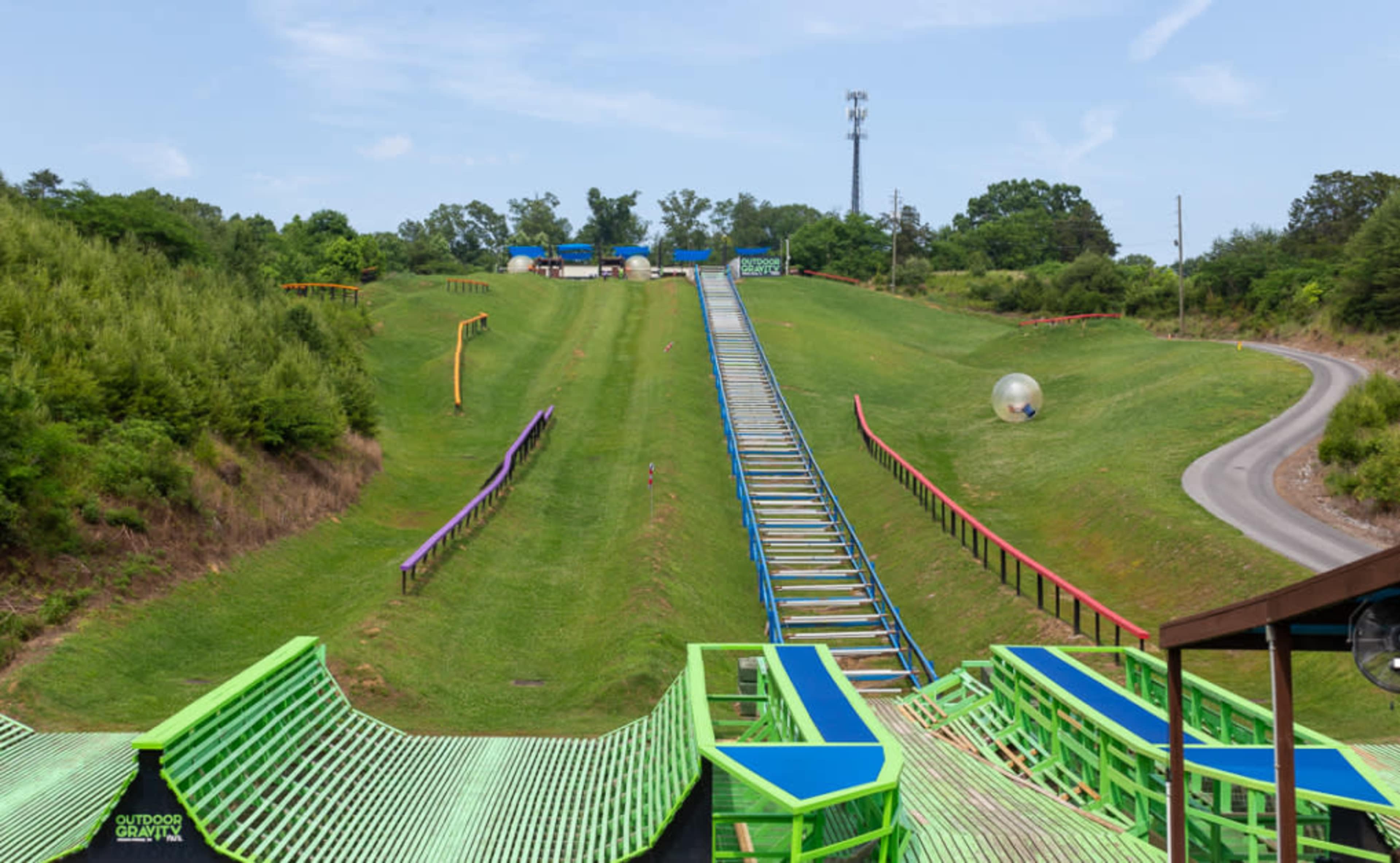 A large green hill with a ski jump ramp and a slide, surrounded by trees and a winding road in the background.
