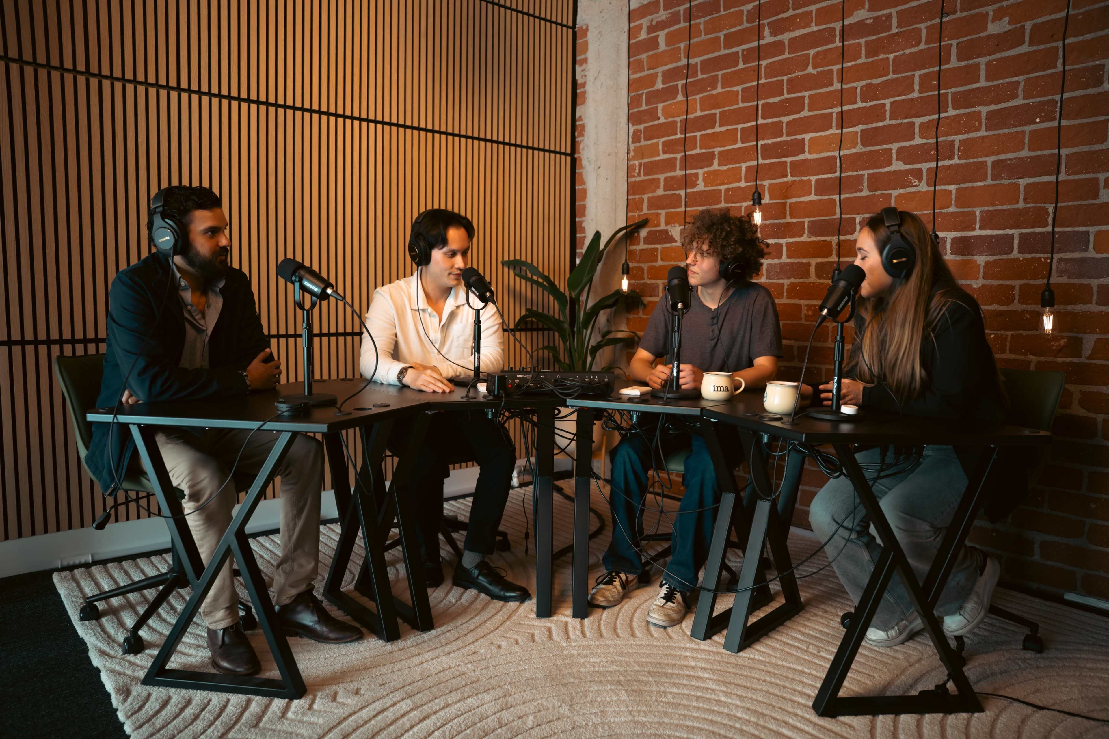 Four people sit at a table in a well-lit room with exposed brick walls, engaged in a podcast discussion, each using a microphone.