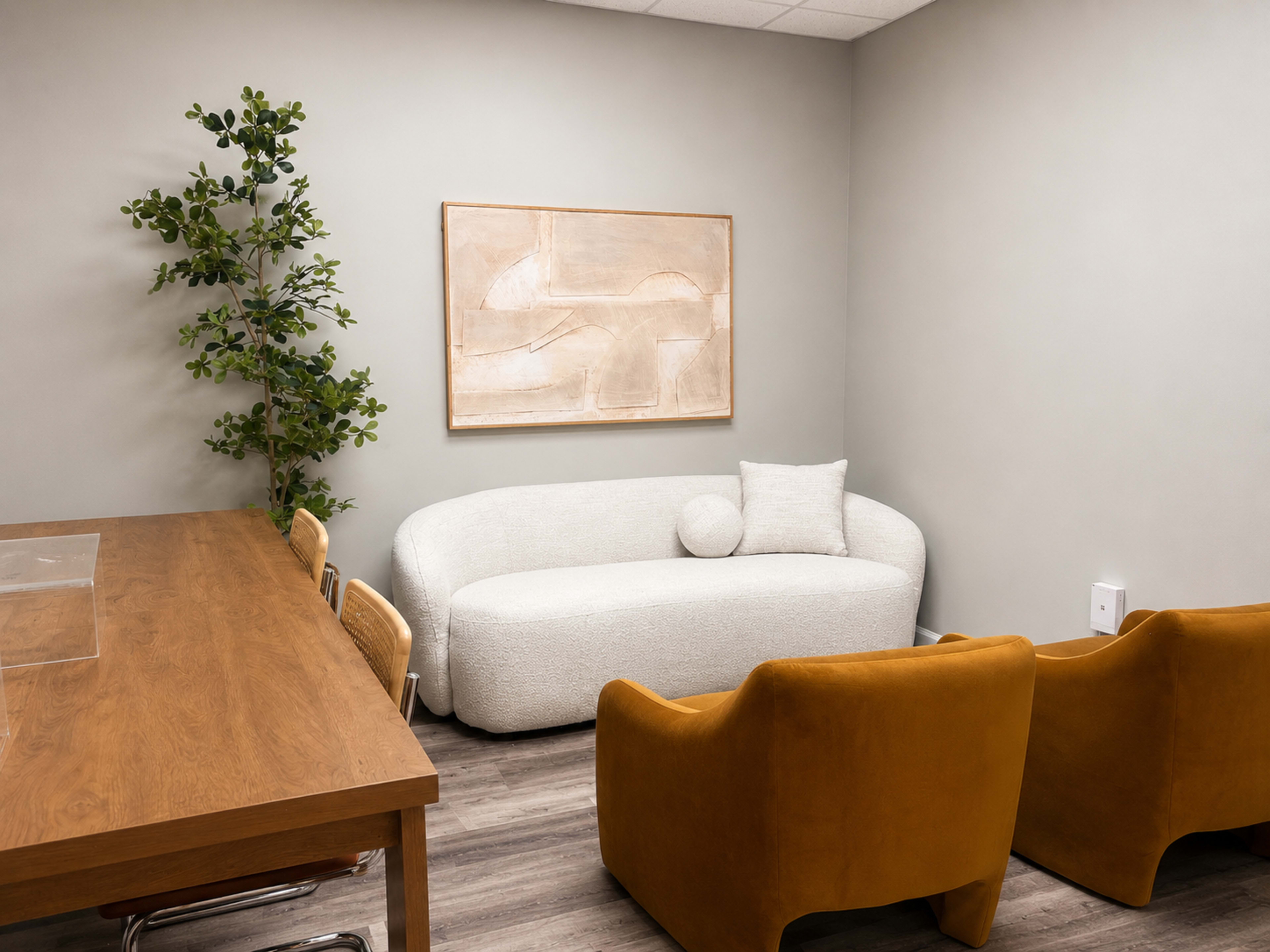 The image shows a small, modern room with a curved white sofa, two mustard-colored armchairs, a wooden table, and a decorative plant against a light-colored wall.