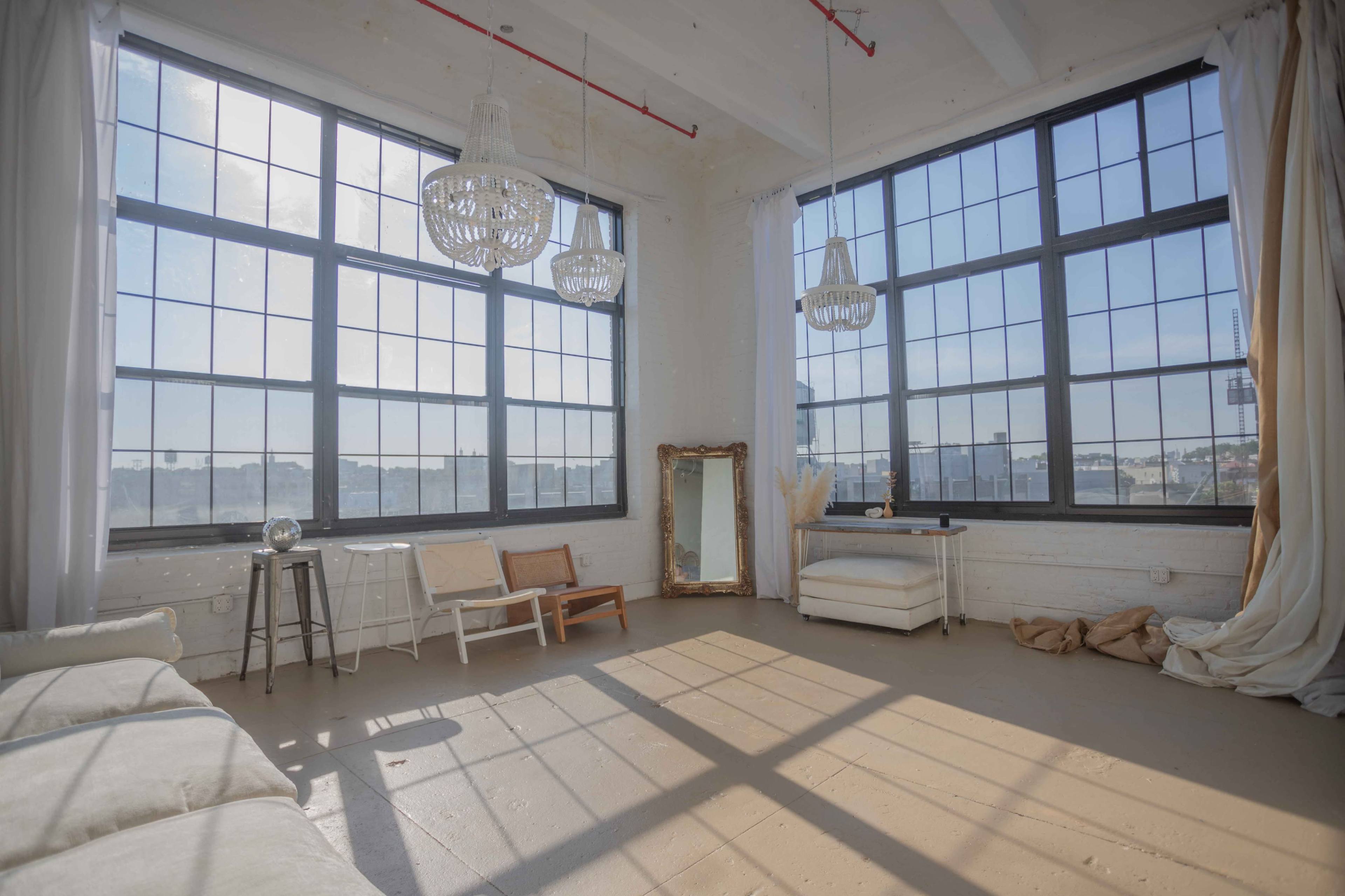 A spacious, bright room with large windows, a hardwood floor, and minimal furniture including a sofa, a mirror, and a small table.