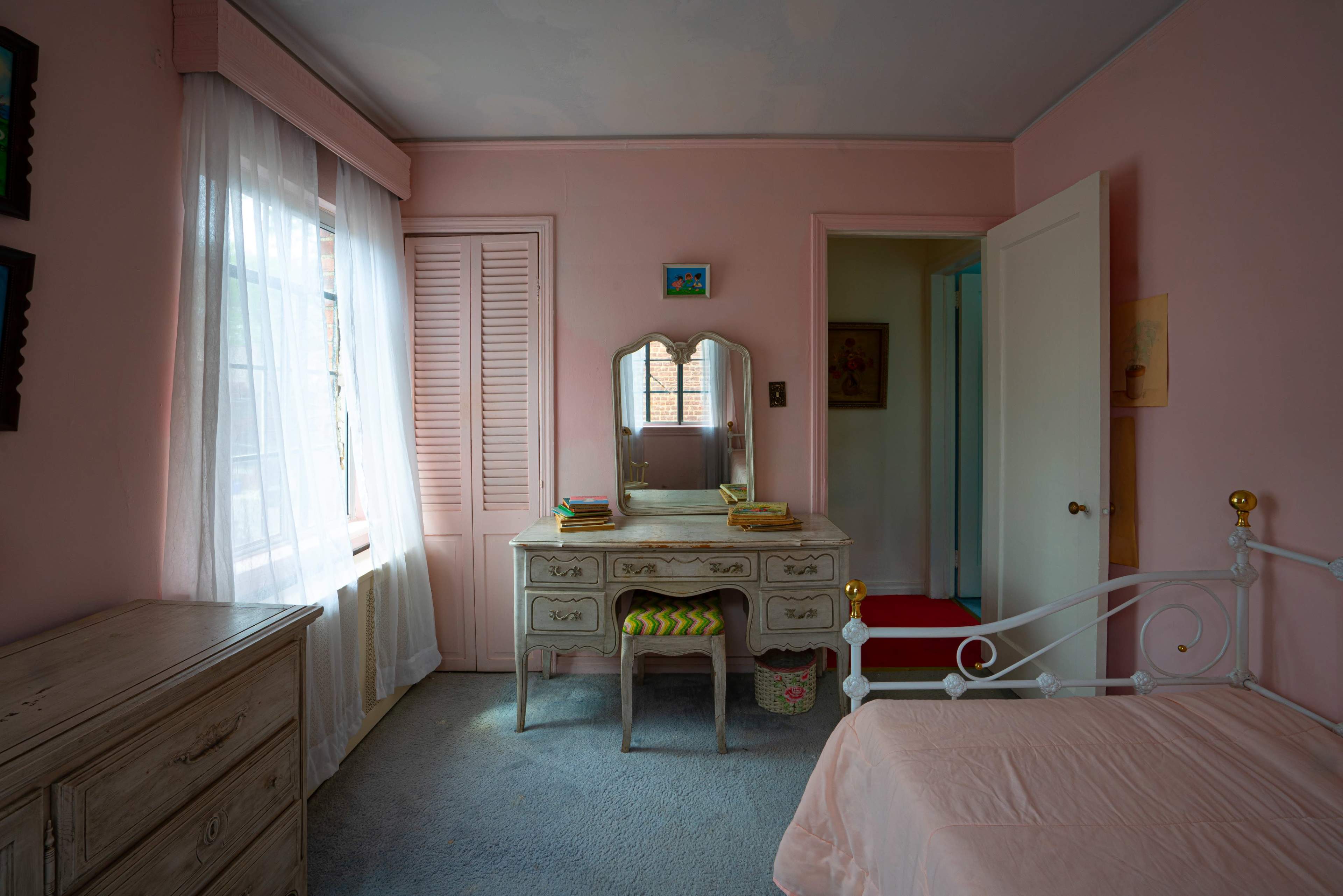The room features pink walls, a white bed, a vintage dresser with a mirror, and a large window dressed with sheer curtains.