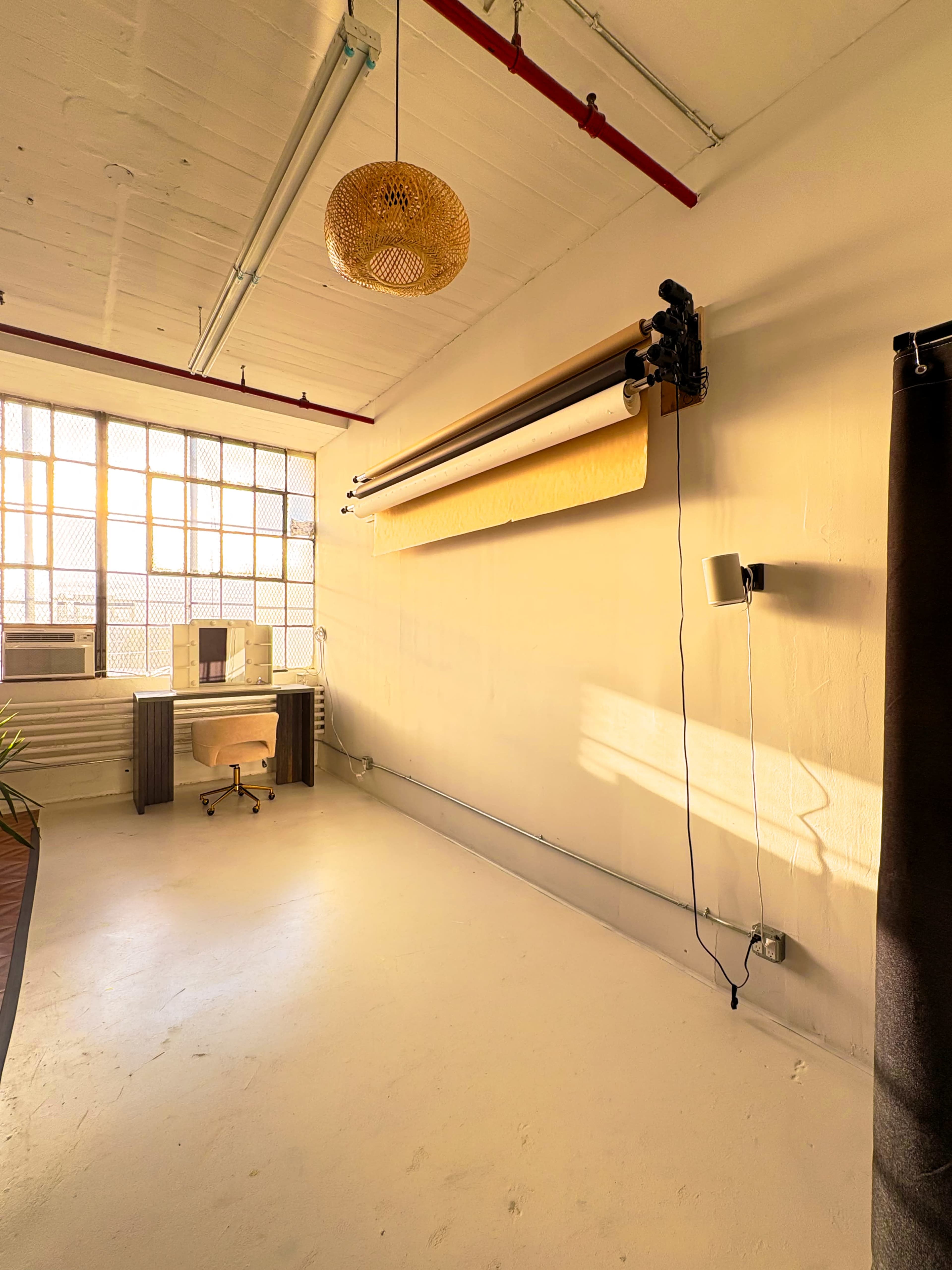 The image shows an empty, well-lit studio space with a large window, overhead paper roll backdrop, and minimal furniture including a desk and two black curtains.