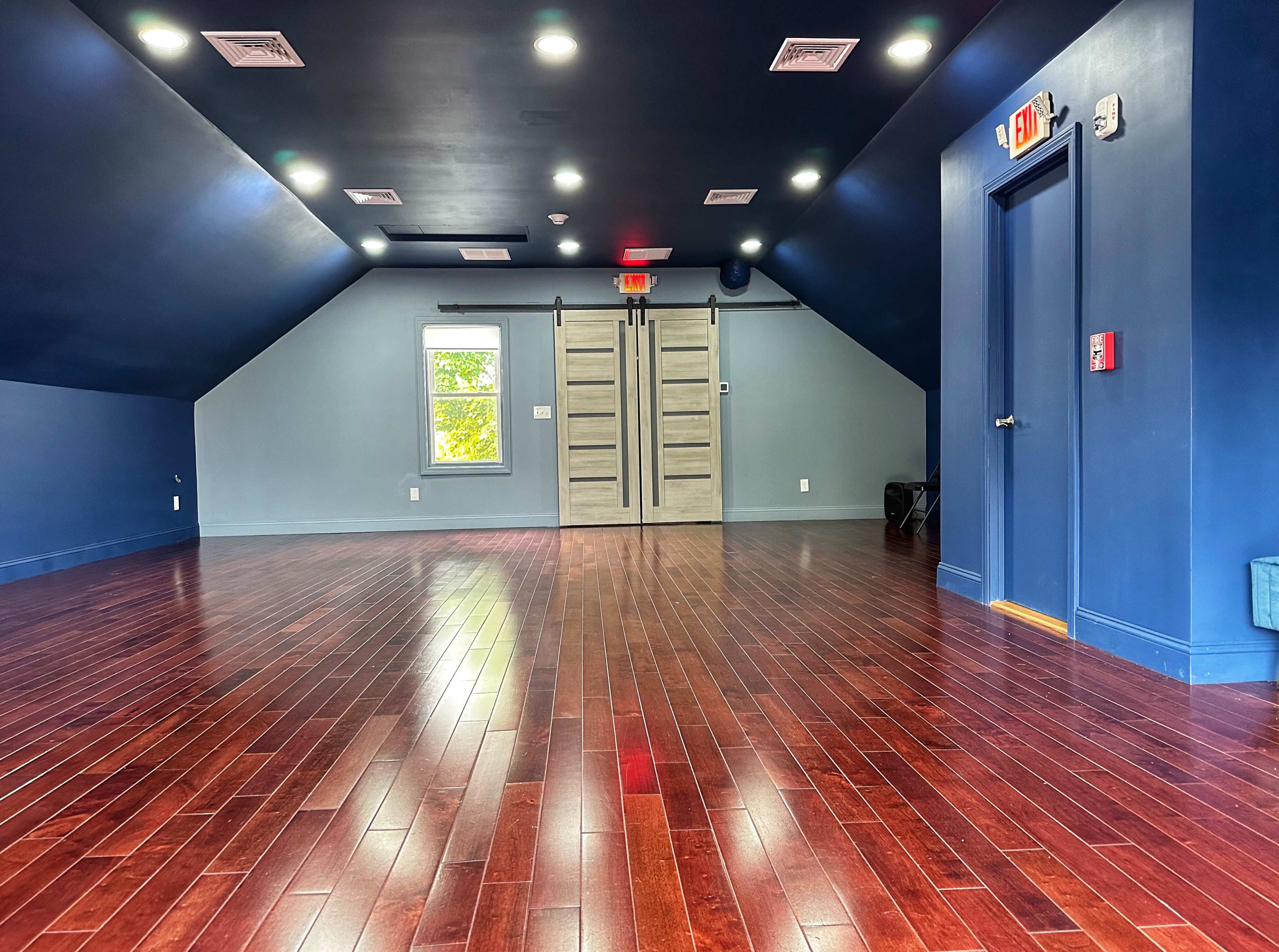 The image shows a spacious, well-lit room with dark wooden flooring, blue walls, and a set of double doors on one side.