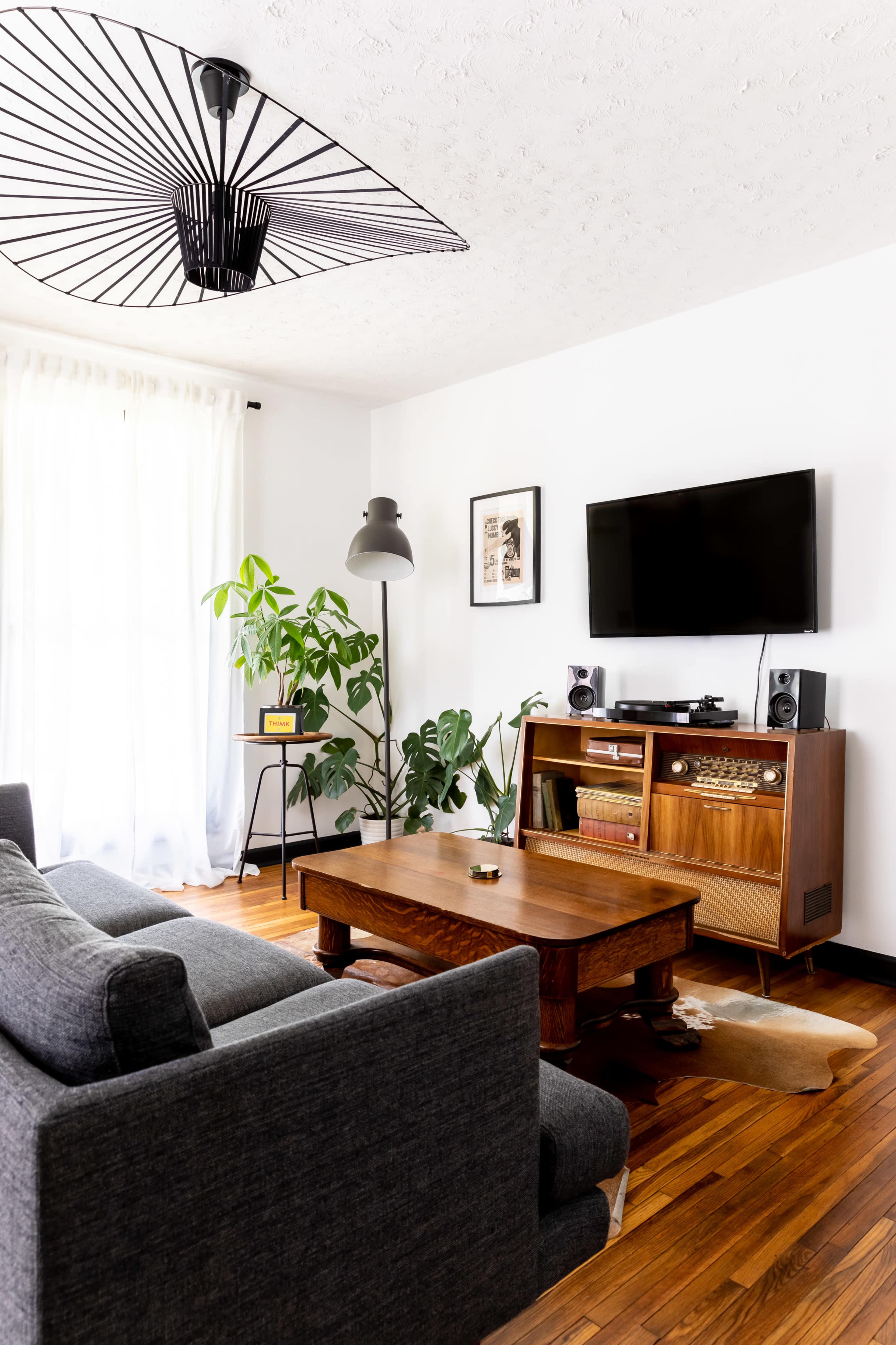 A modern living room features a gray sofa, wooden coffee table, floor lamp, and entertainment unit with a TV, surrounded by plants and bright natural light.