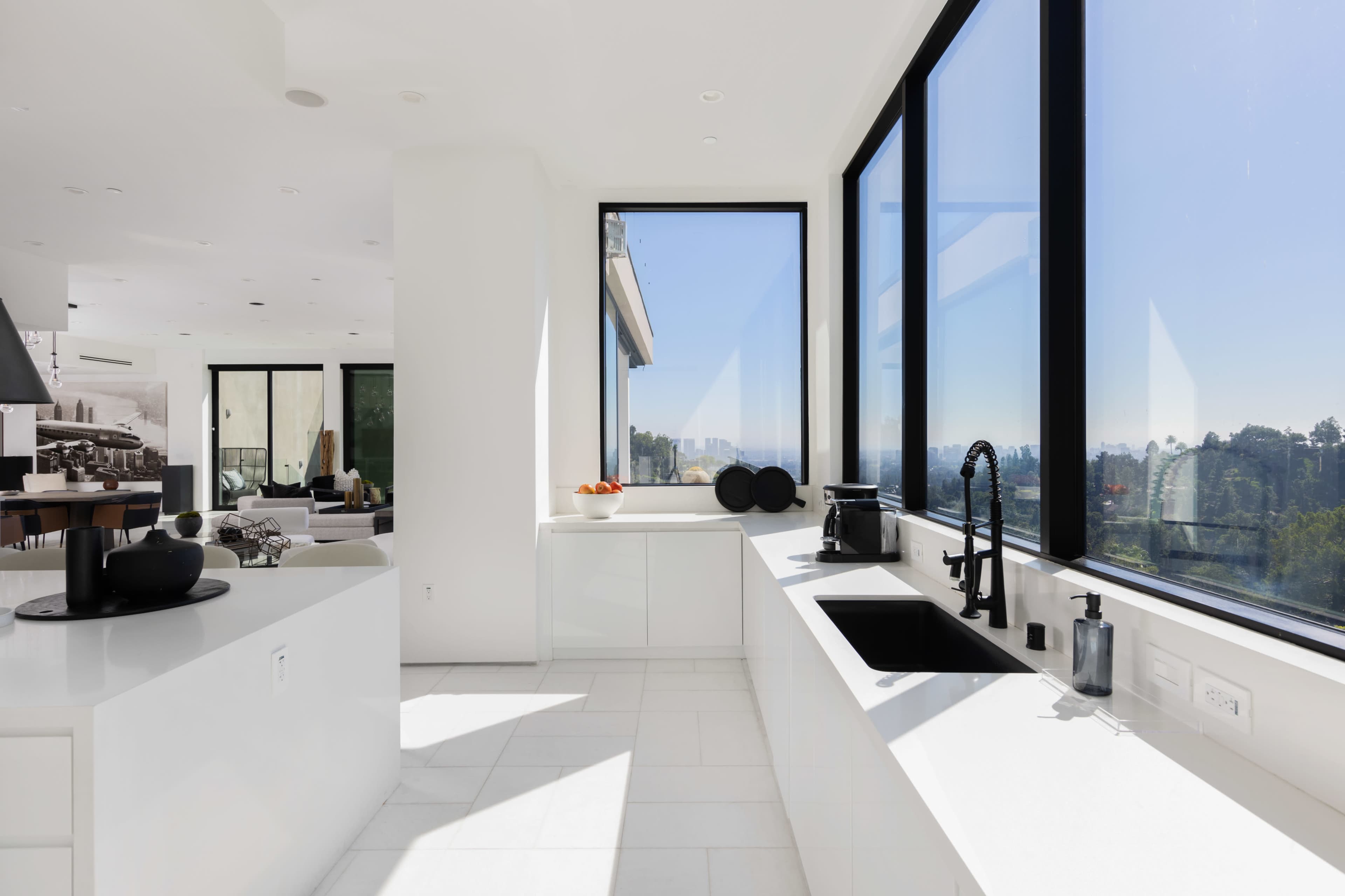 The kitchen features large windows with a view of the landscape, showcasing a minimalist design with white countertops and cabinetry.