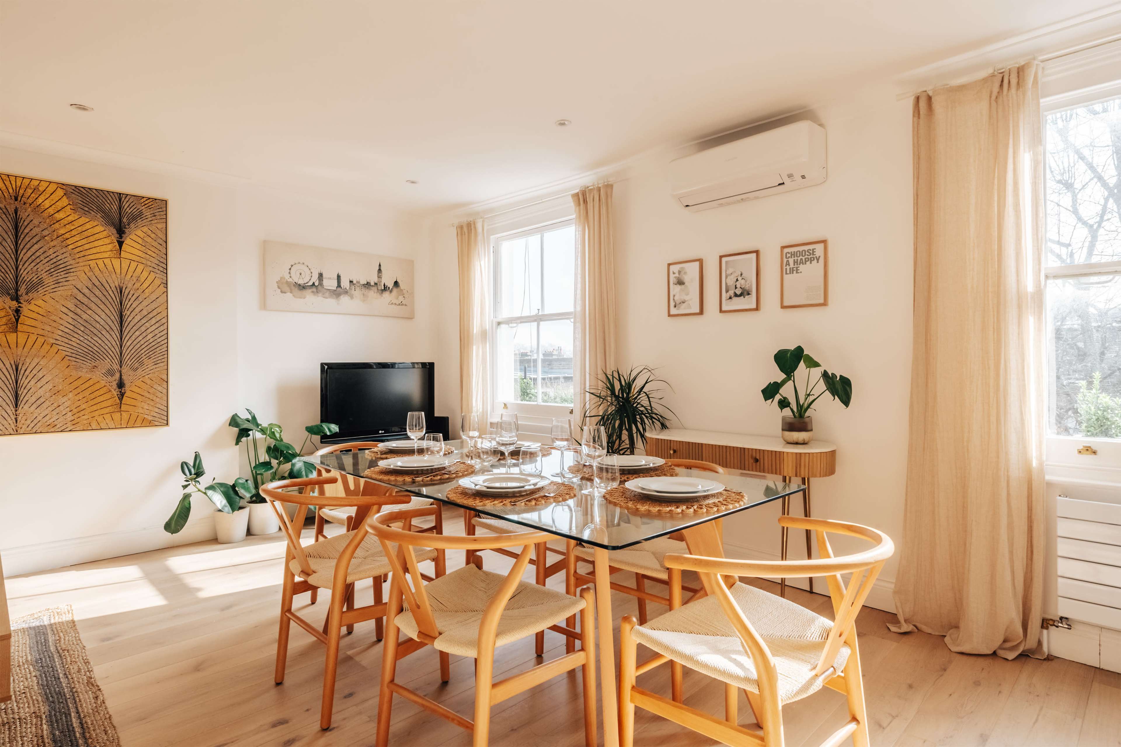 A dining room features a glass table set for dinner, surrounded by wooden chairs, with plants and artwork adorning the walls.