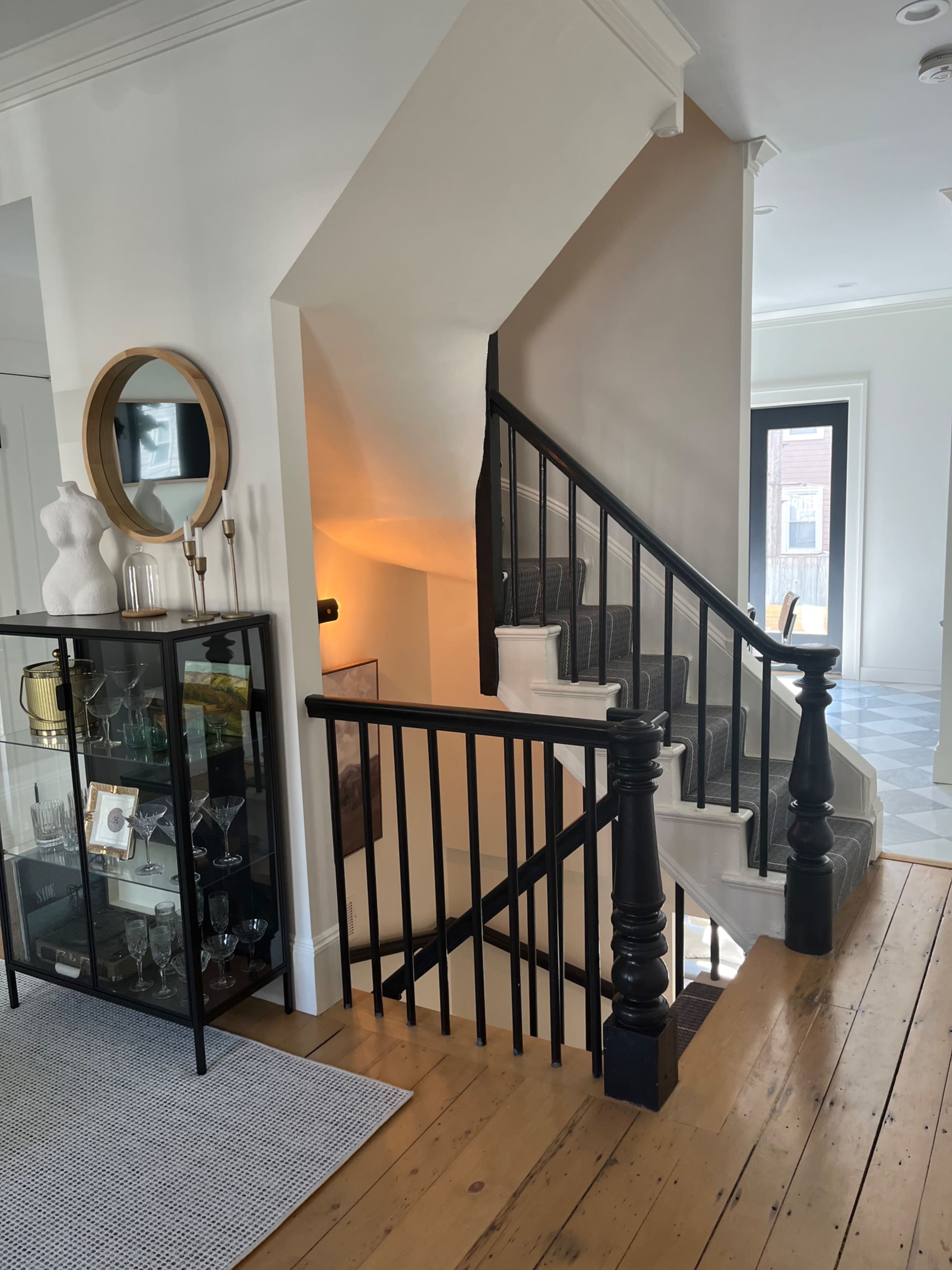 A staircase with black railings ascends to a second floor in a bright, modern entryway featuring a glass display cabinet and a round mirror on the wall.