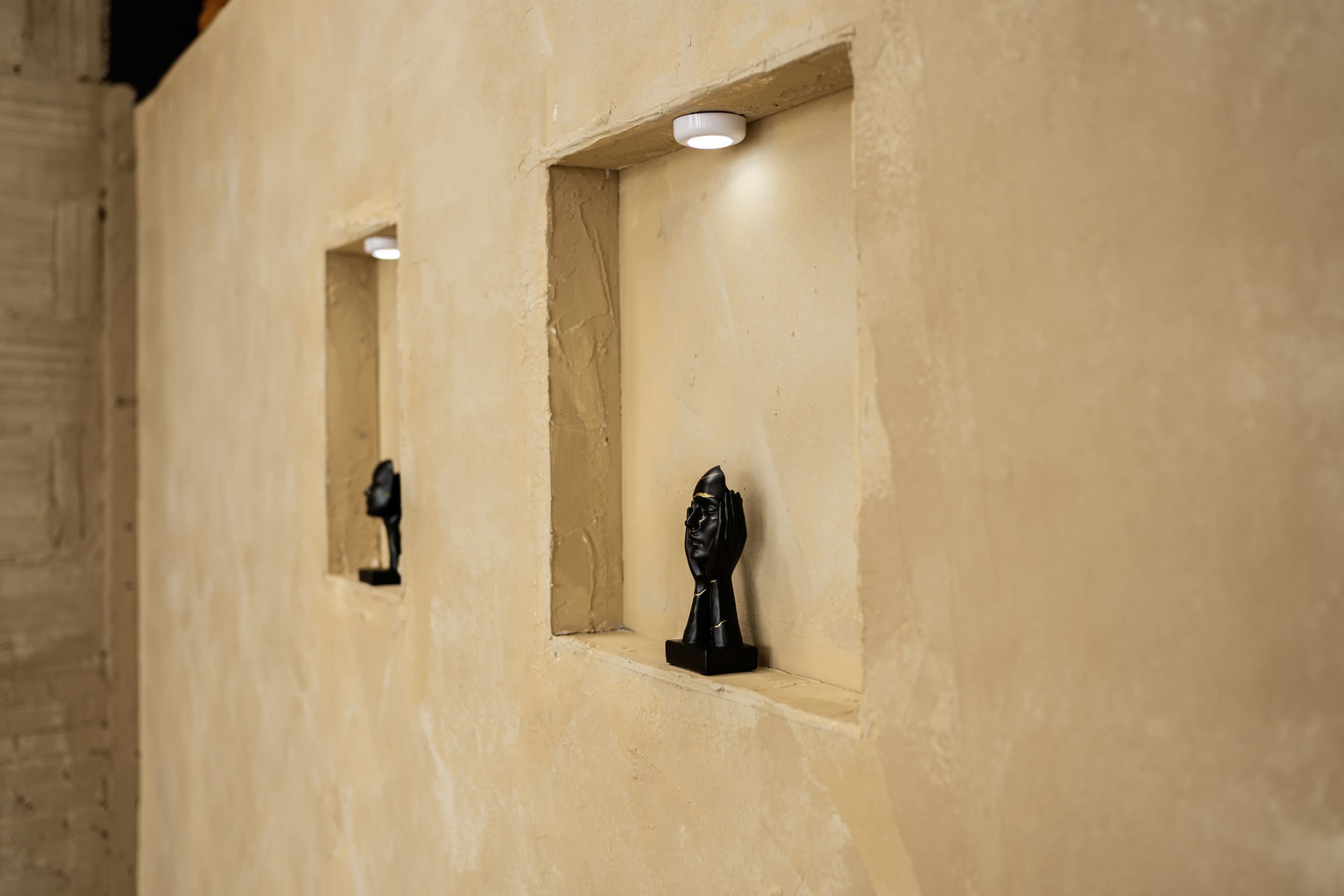 Two small black busts are displayed in recessed niches on a textured beige wall, illuminated by overhead lights.