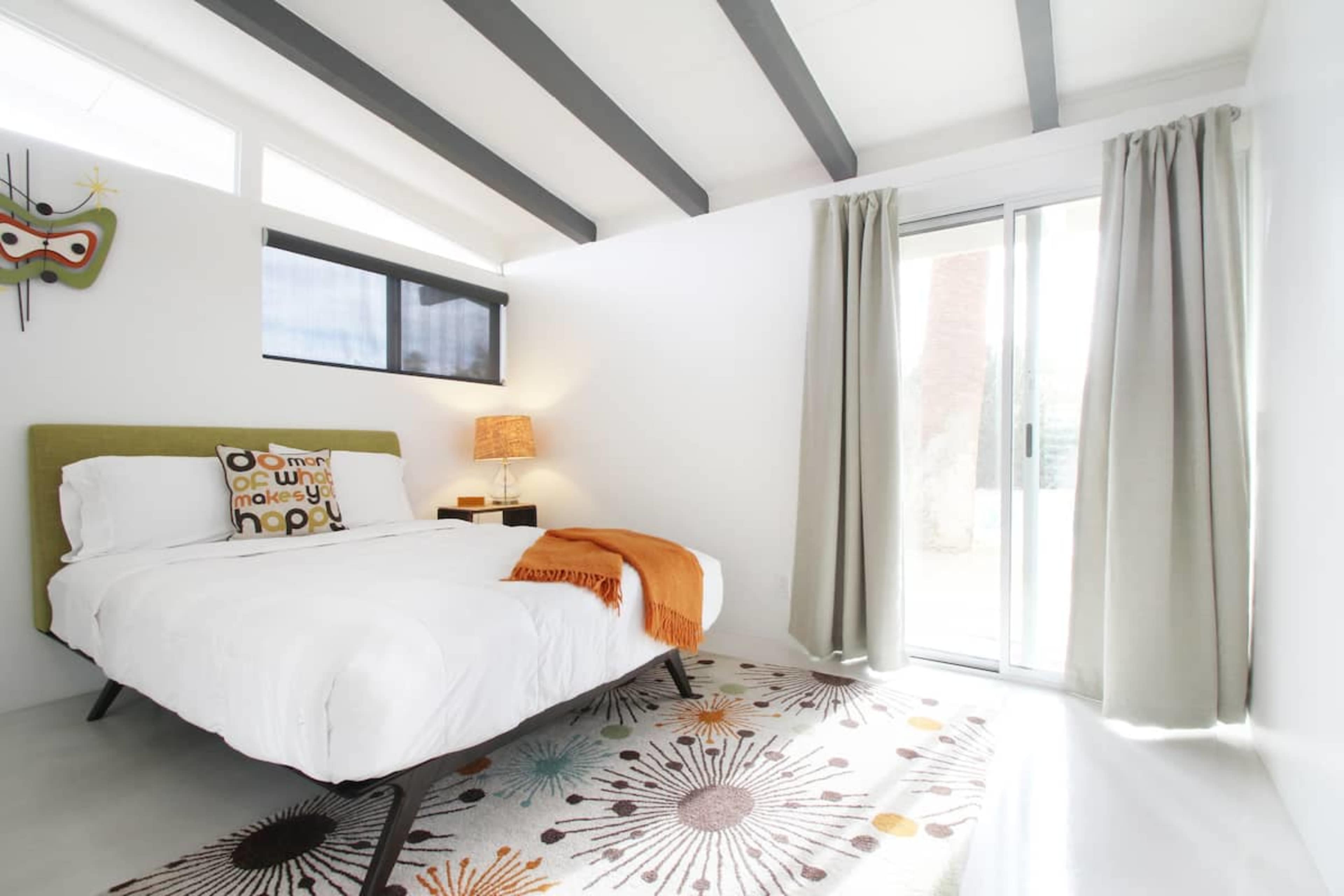 A modern bedroom features a bed with a colorful accent pillow, a decorative lamp, and a sliding glass door that leads outside.