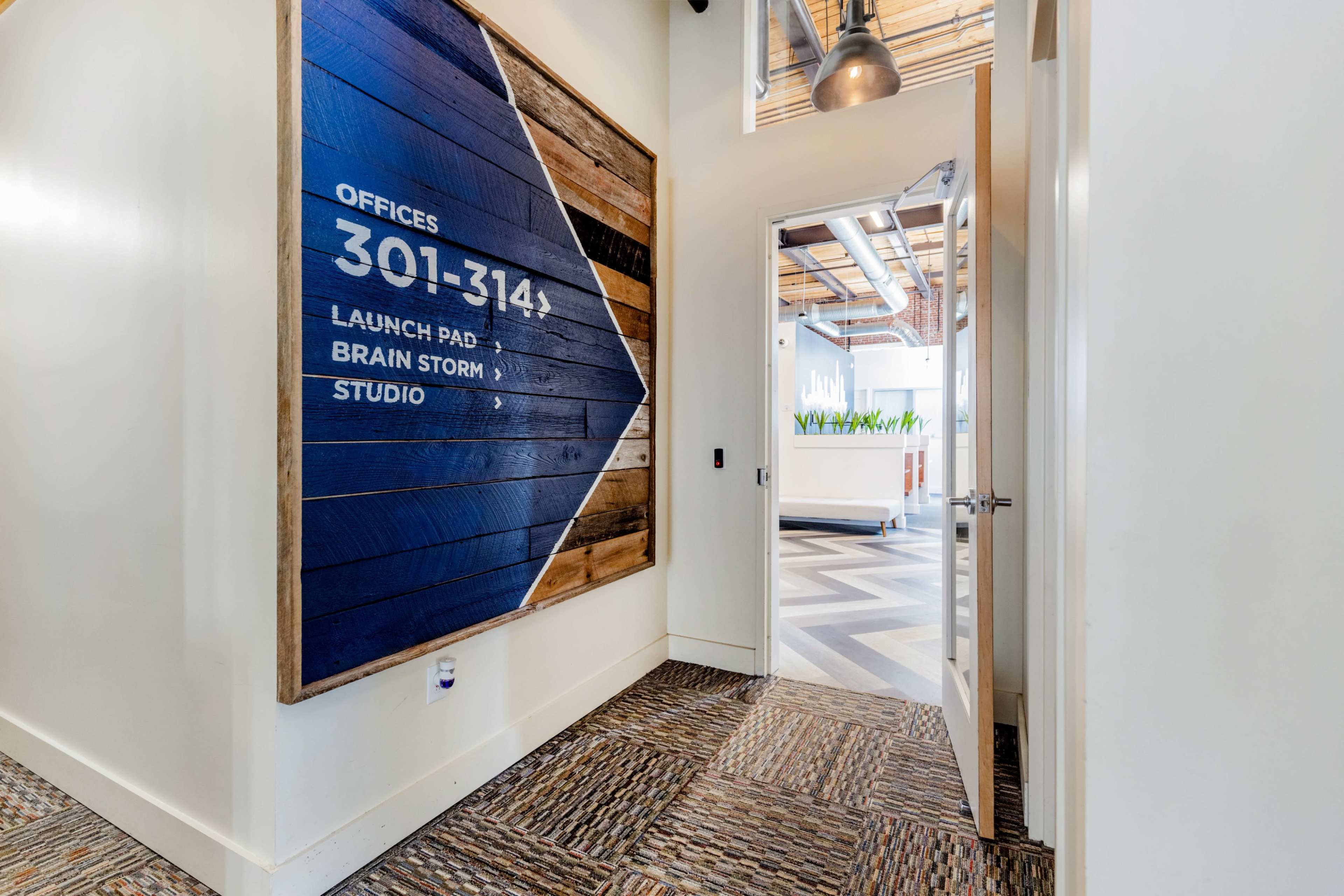 A hallway features a wooden sign with room numbers and labels, leading to an open workspace with green plants visible.