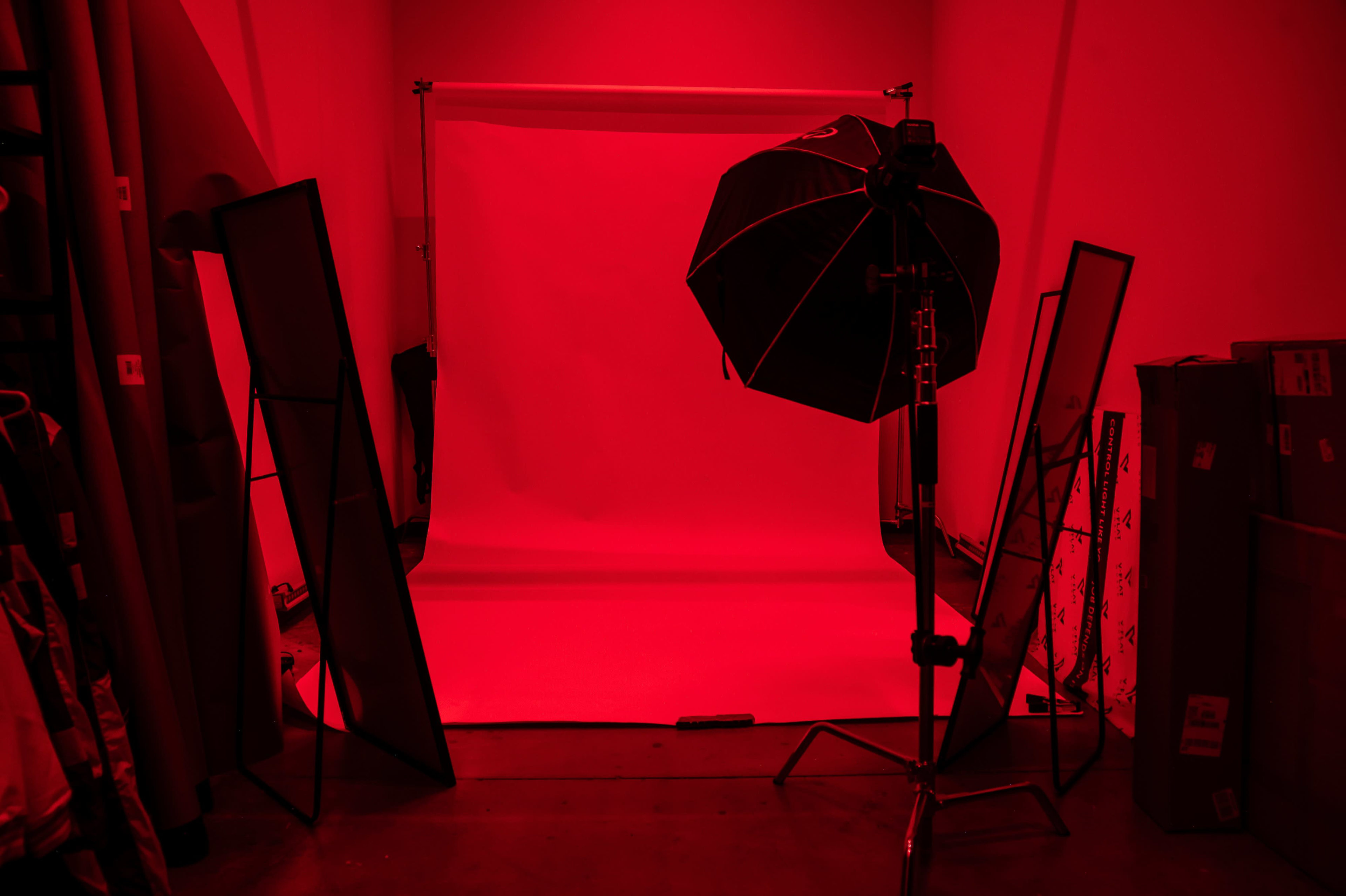 A photography studio features a red backdrop illuminated by an umbrella light, surrounded by mirrors and equipment.