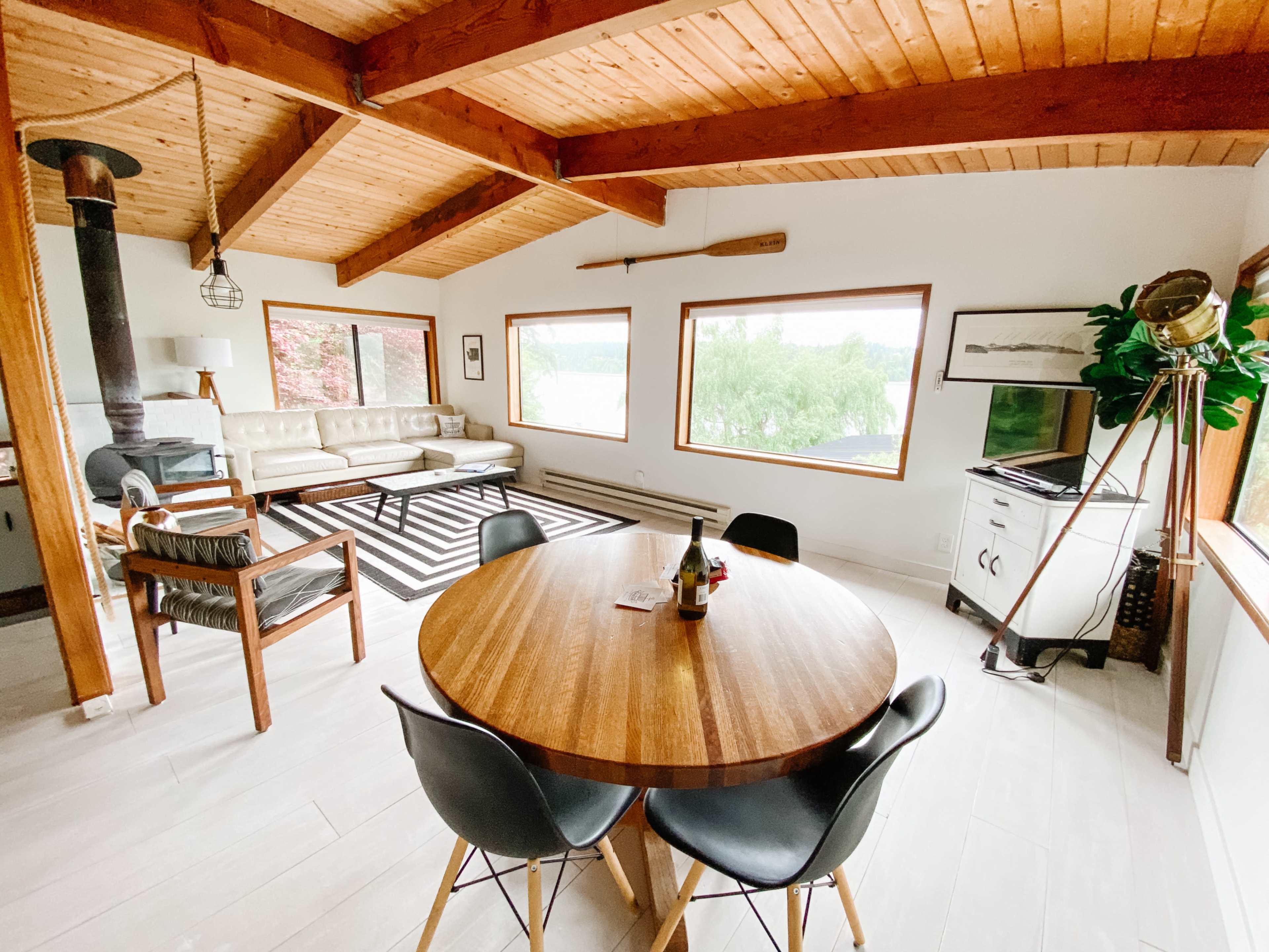 A modern living space featuring a round wooden dining table, black chairs, a contemporary sofa, large windows with views of greenery, and a wood stove.