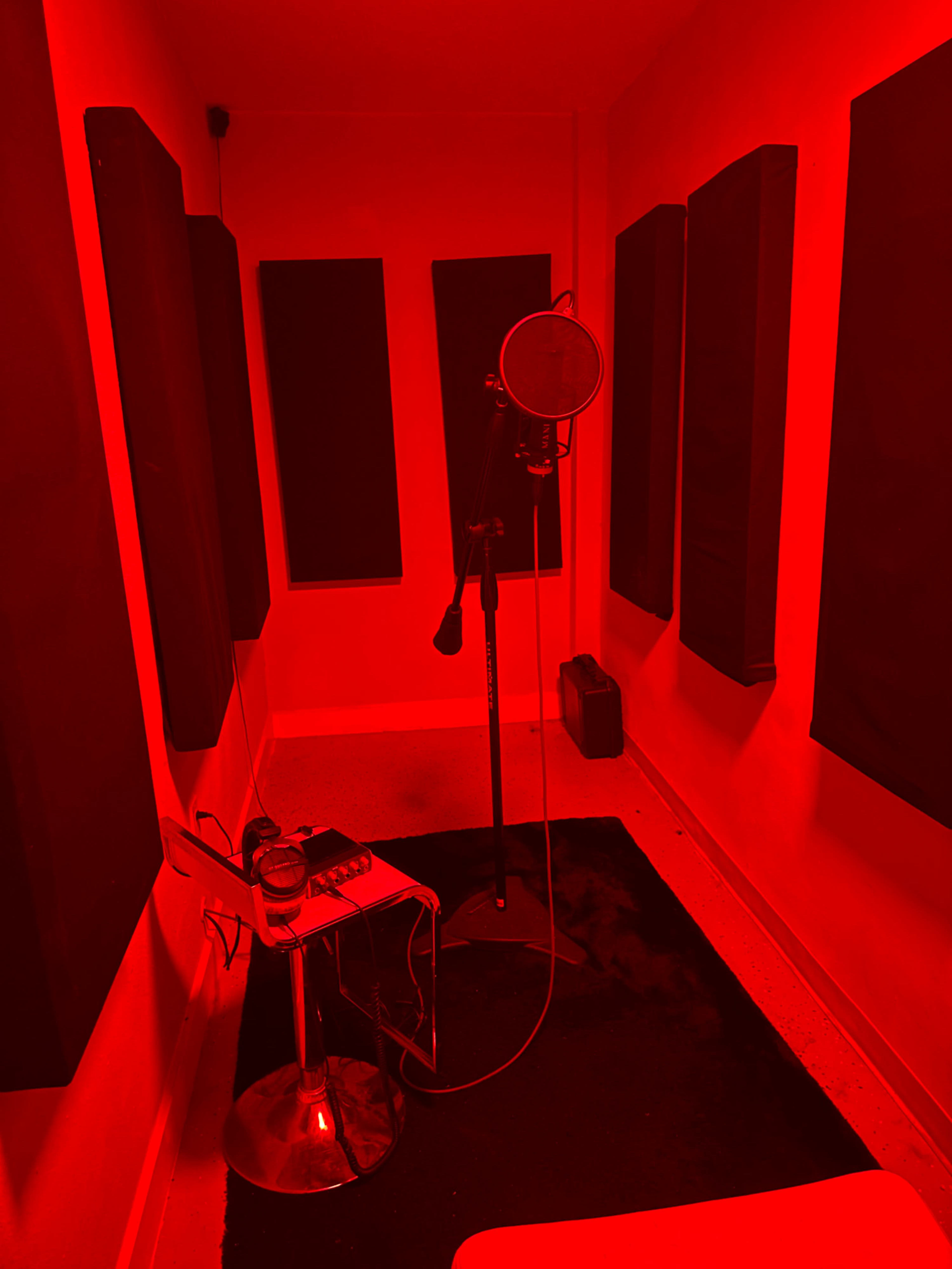 A small recording studio with red lighting, featuring a microphone on a stand, a stool, and soundproof panels on the walls.