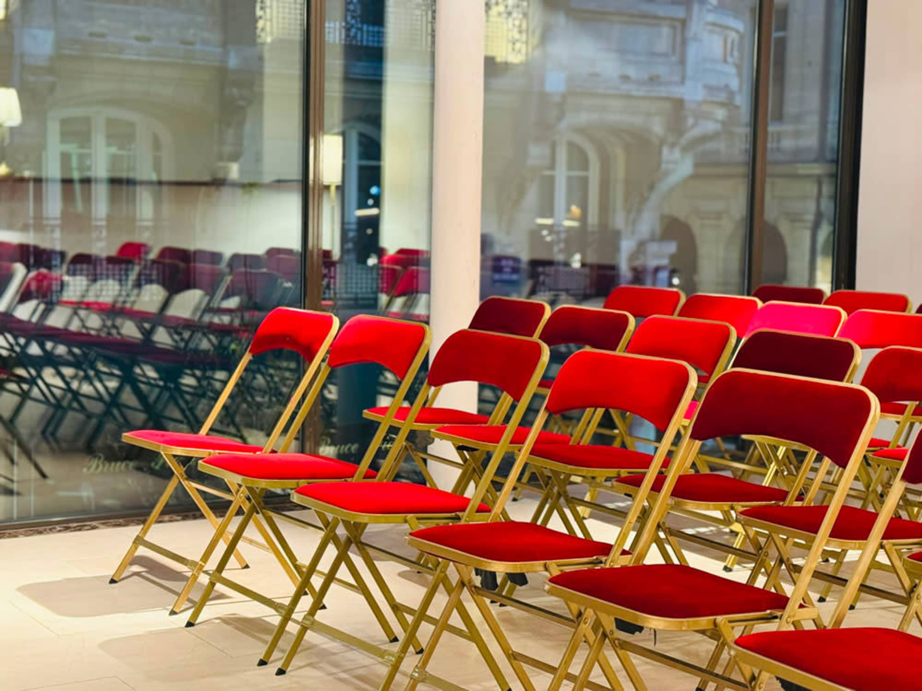 A set of red folding chairs with gold frames is arranged in rows facing a large window.