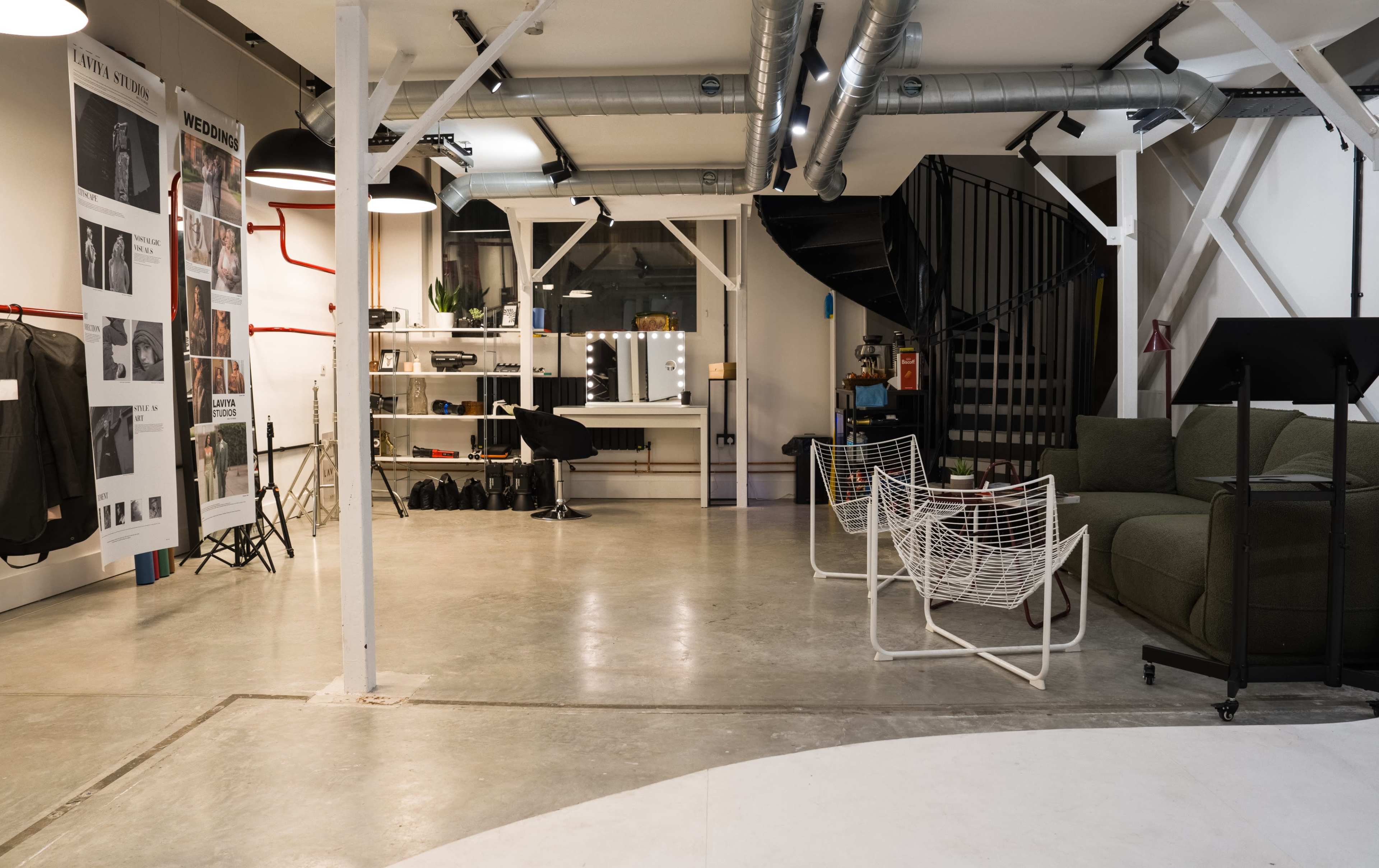 The image shows a modern, industrial-style studio or workspace with gray concrete flooring, a seating area, and a decorative wall featuring various mounted items.