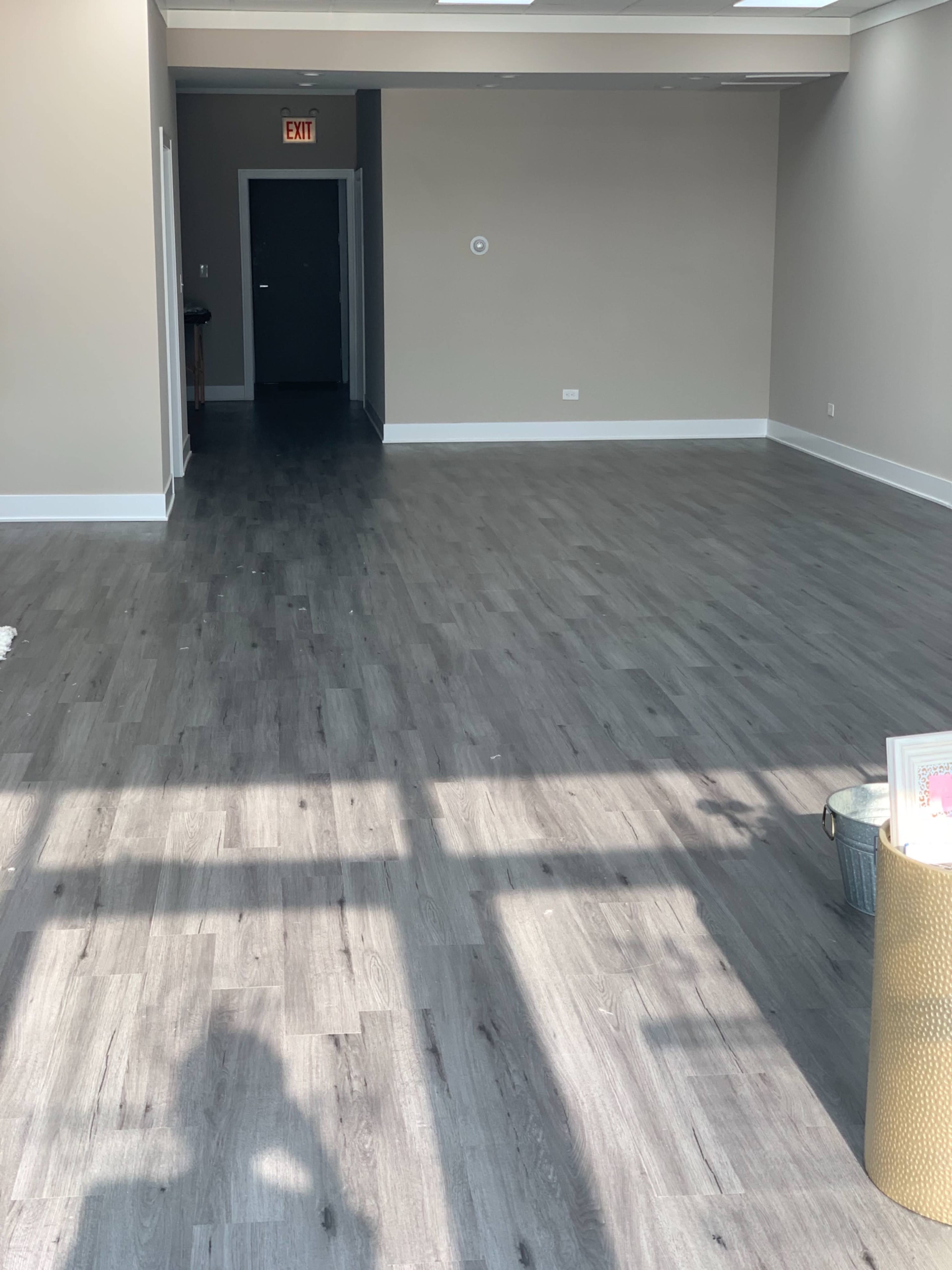 A spacious, unfurnished room with gray laminate flooring and light-colored walls, featuring a doorway at the far end.