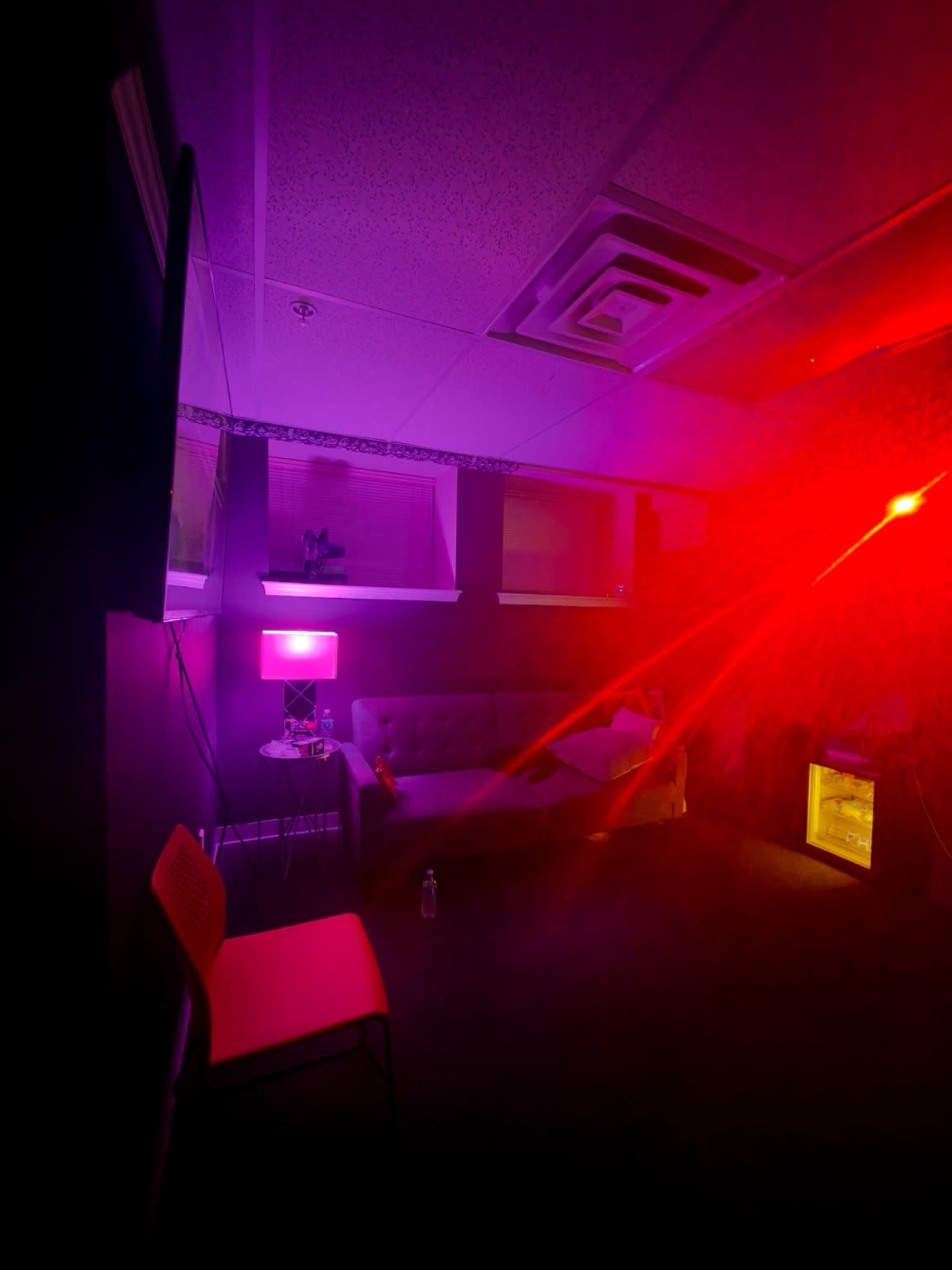 A dimly lit room features a gray sofa, a pink lamp, and a red light creating a vibrant atmosphere.