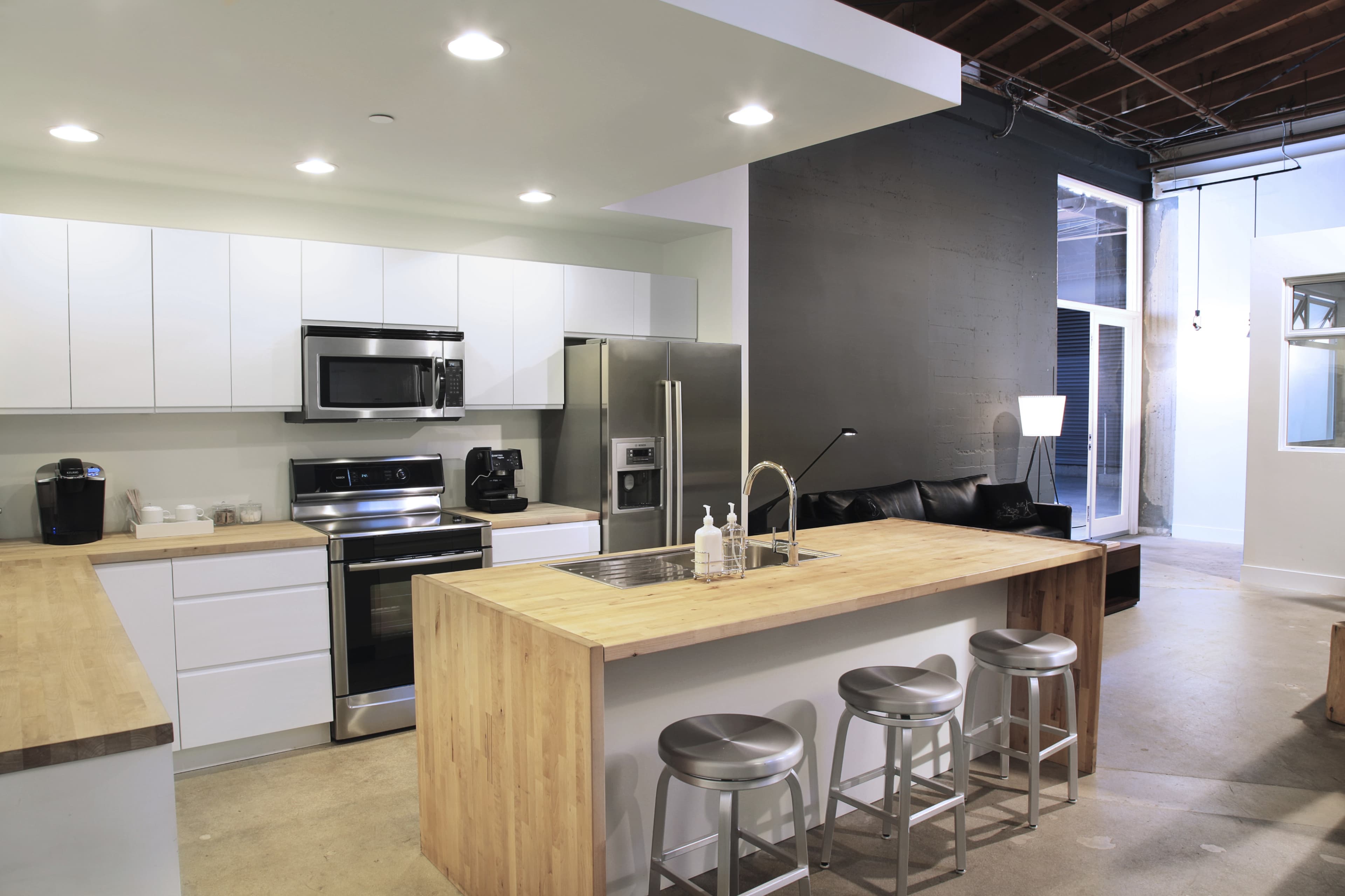 The image shows a modern kitchen with white cabinetry, stainless steel appliances, and a large wooden island with three metal stools.